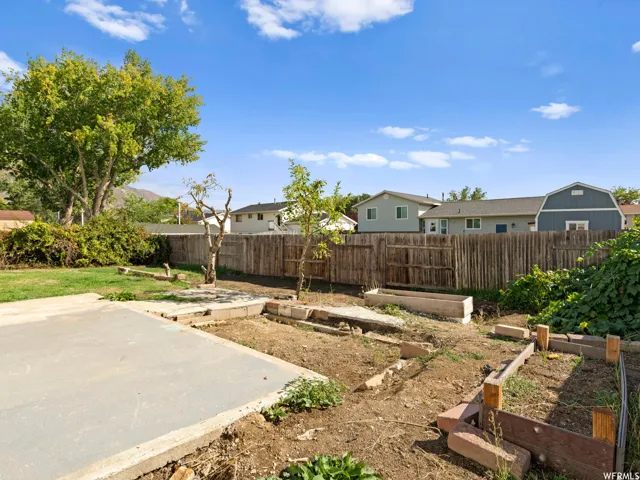 Spacious backyard featuring a large concrete patio, mature trees, a wooden privacy fence, and multiple raised garden beds