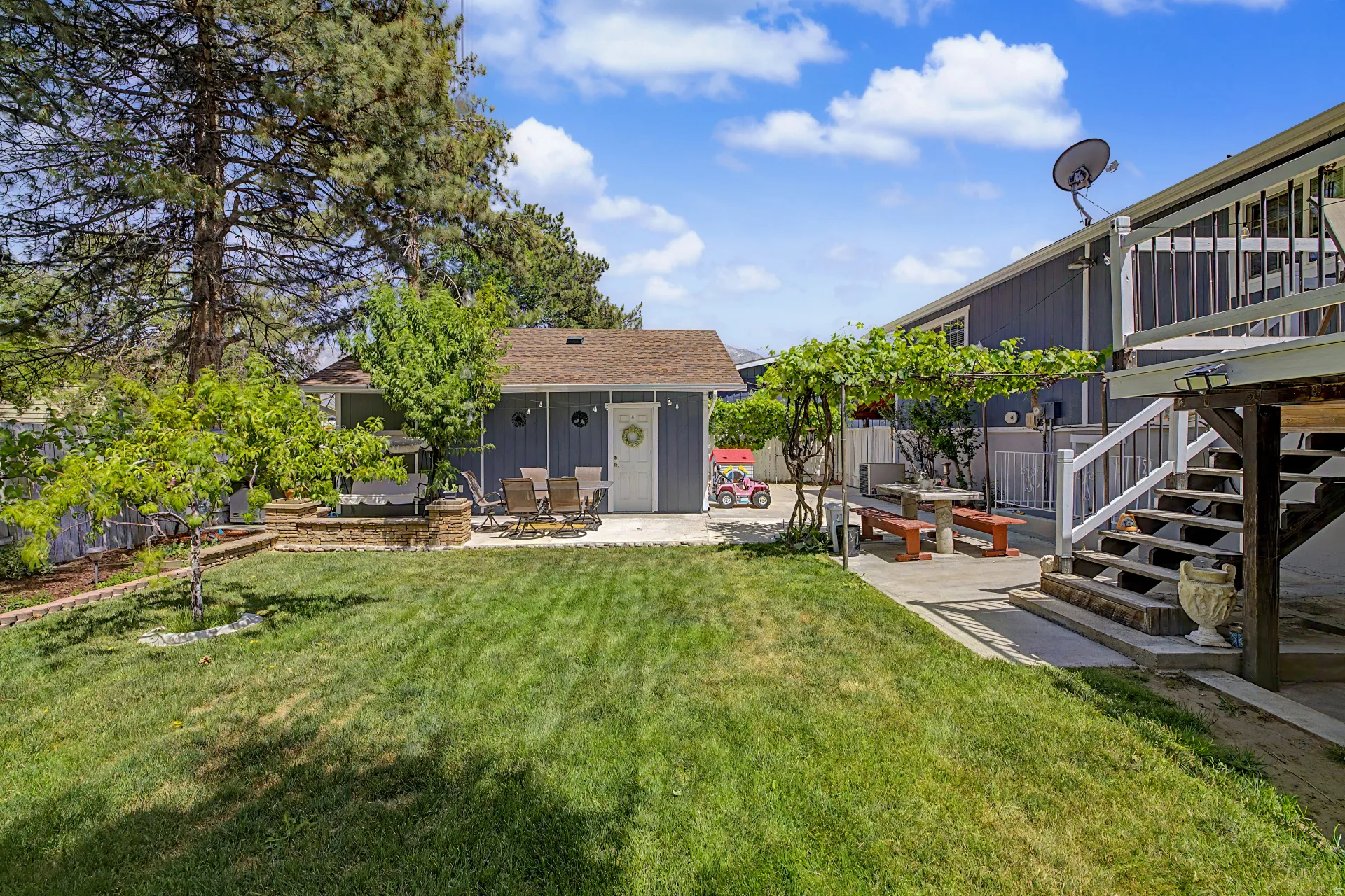 Expansive backyard featuring a lush lawn, mature trees, concrete patio areas, a built-in stone fire pit, and a separate storage shed