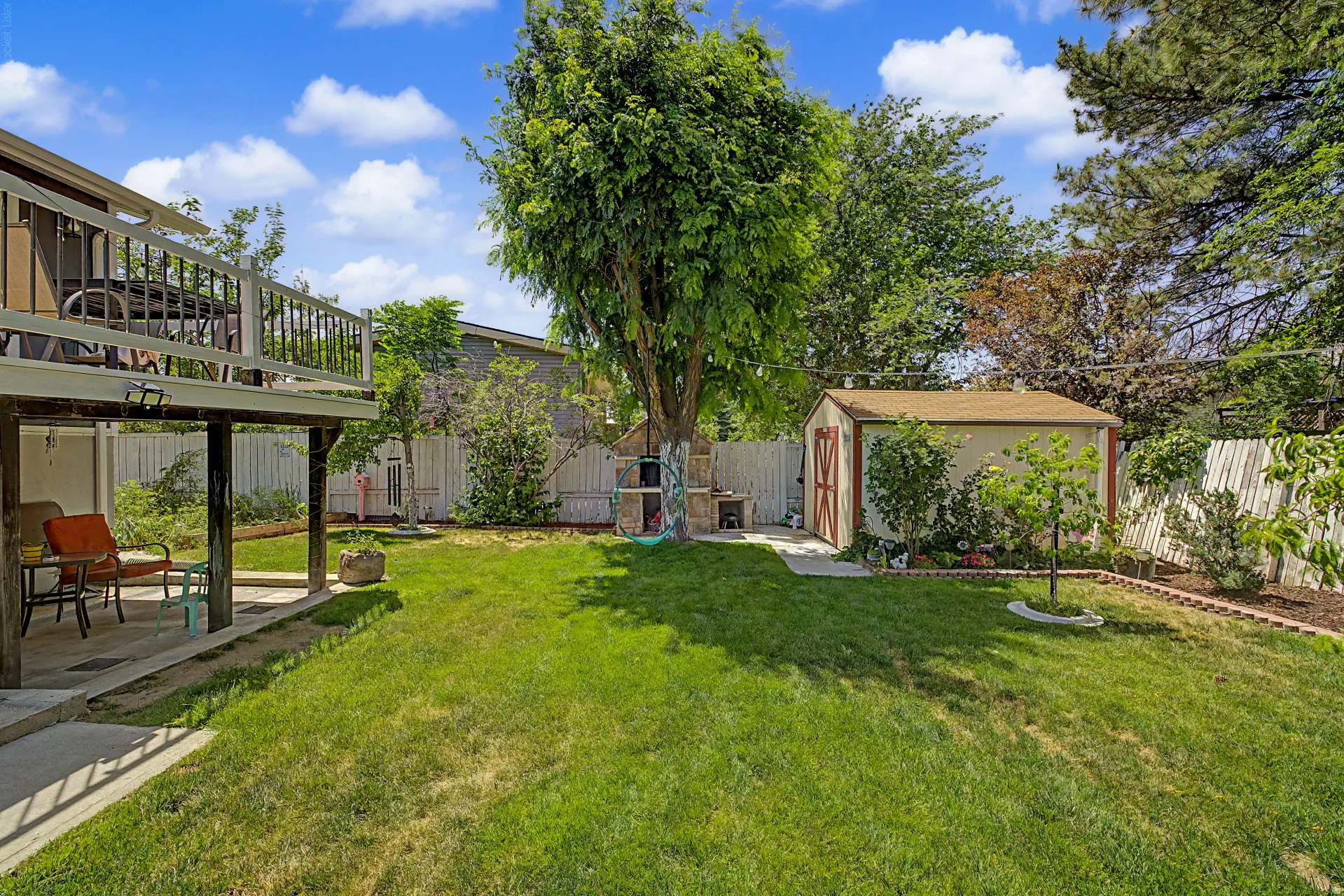 Spacious backyard featuring an elevated deck with white railings, a covered patio area, a detached storage shed, a privacy fence, and mature trees
