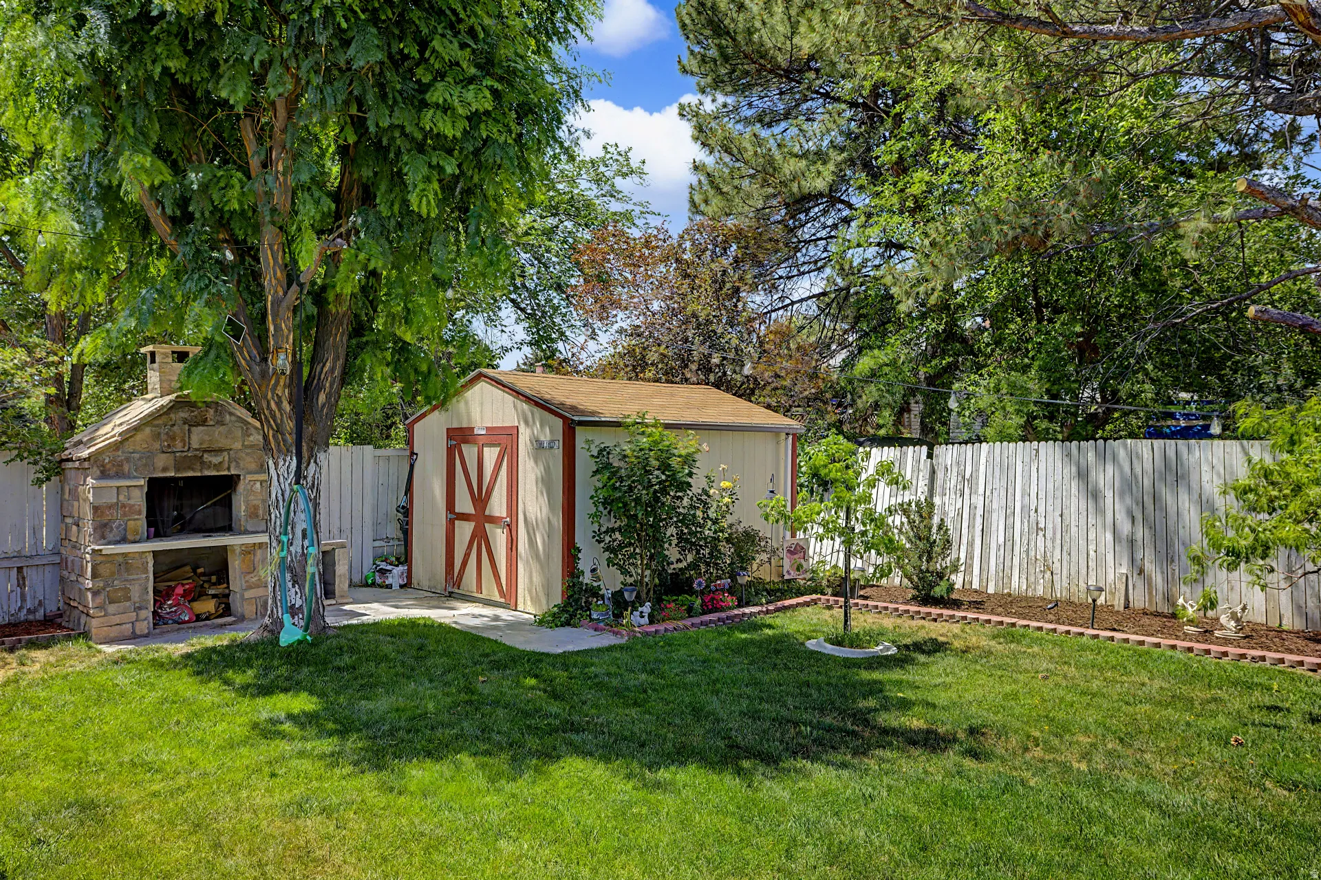 Expansive backyard featuring a manicured lawn, a stone outdoor oven, a storage shed with red-trimmed doors, mature trees, and a perimeter fence