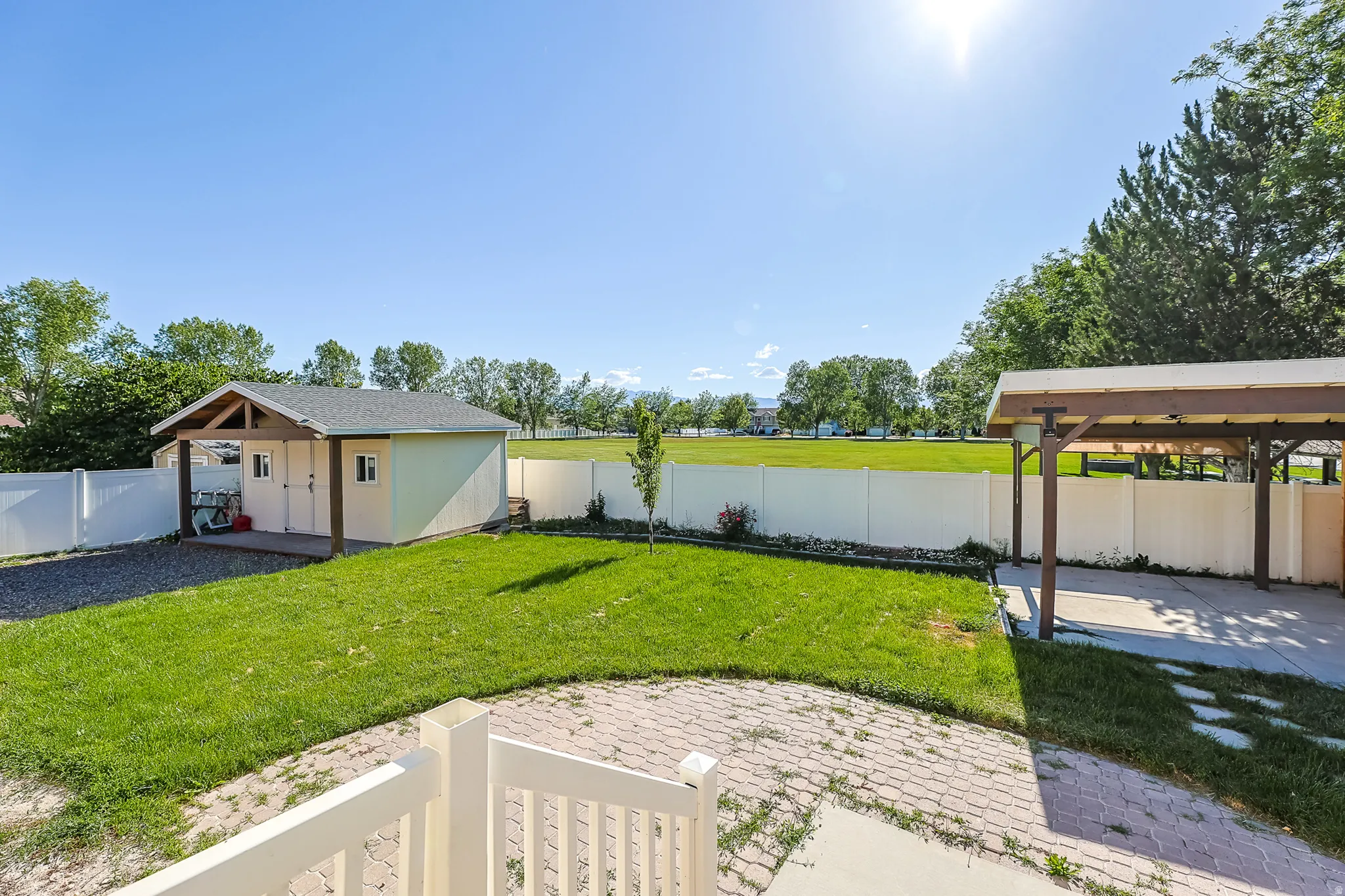 Expansive backyard featuring a manicured lawn, a detached storage shed with an overhang, a covered patio area, and a white privacy fence bordering a large open field