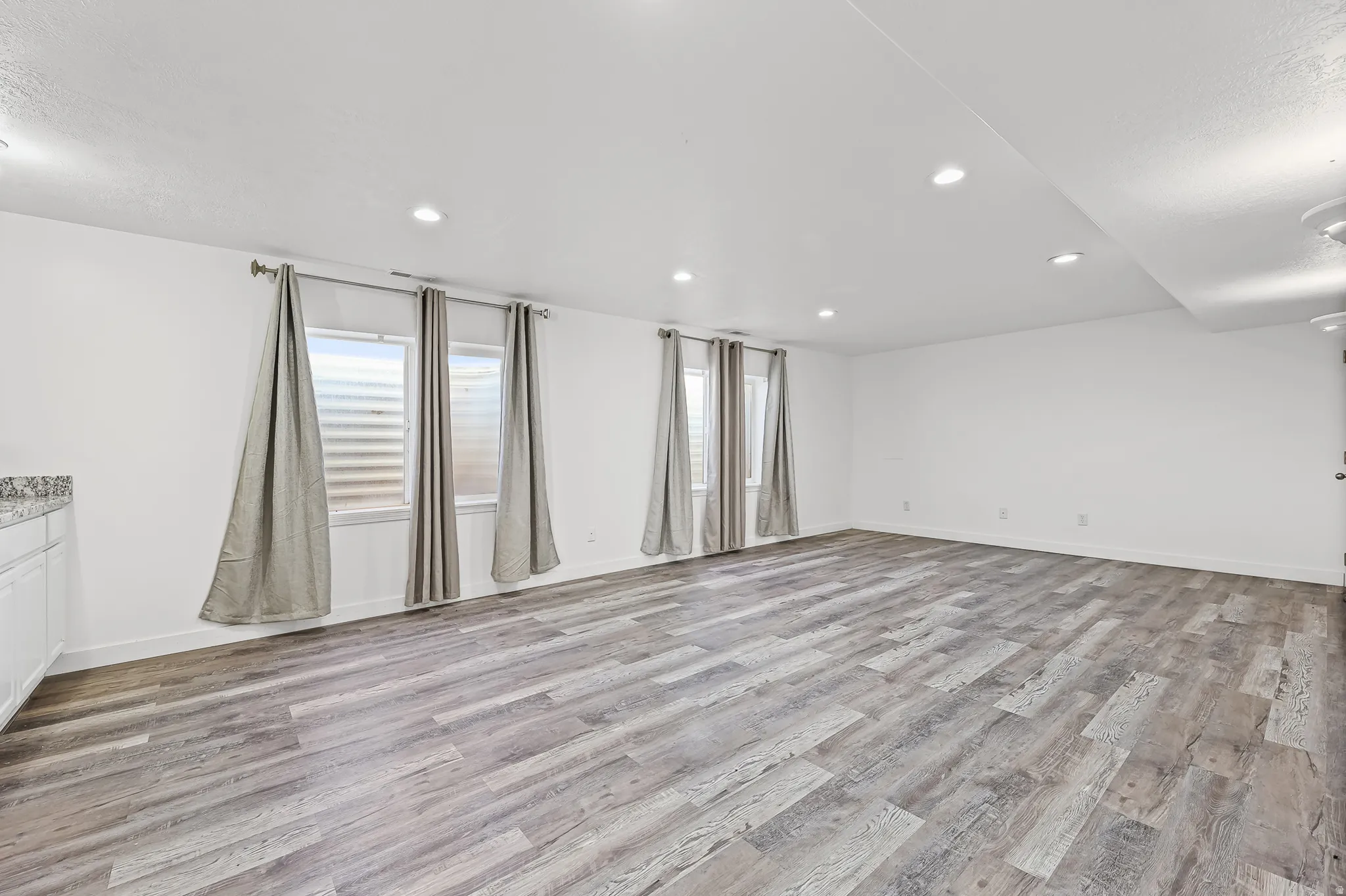 Spacious room featuring wood-finish flooring, white walls, recessed lighting, and multiple windows with blinds