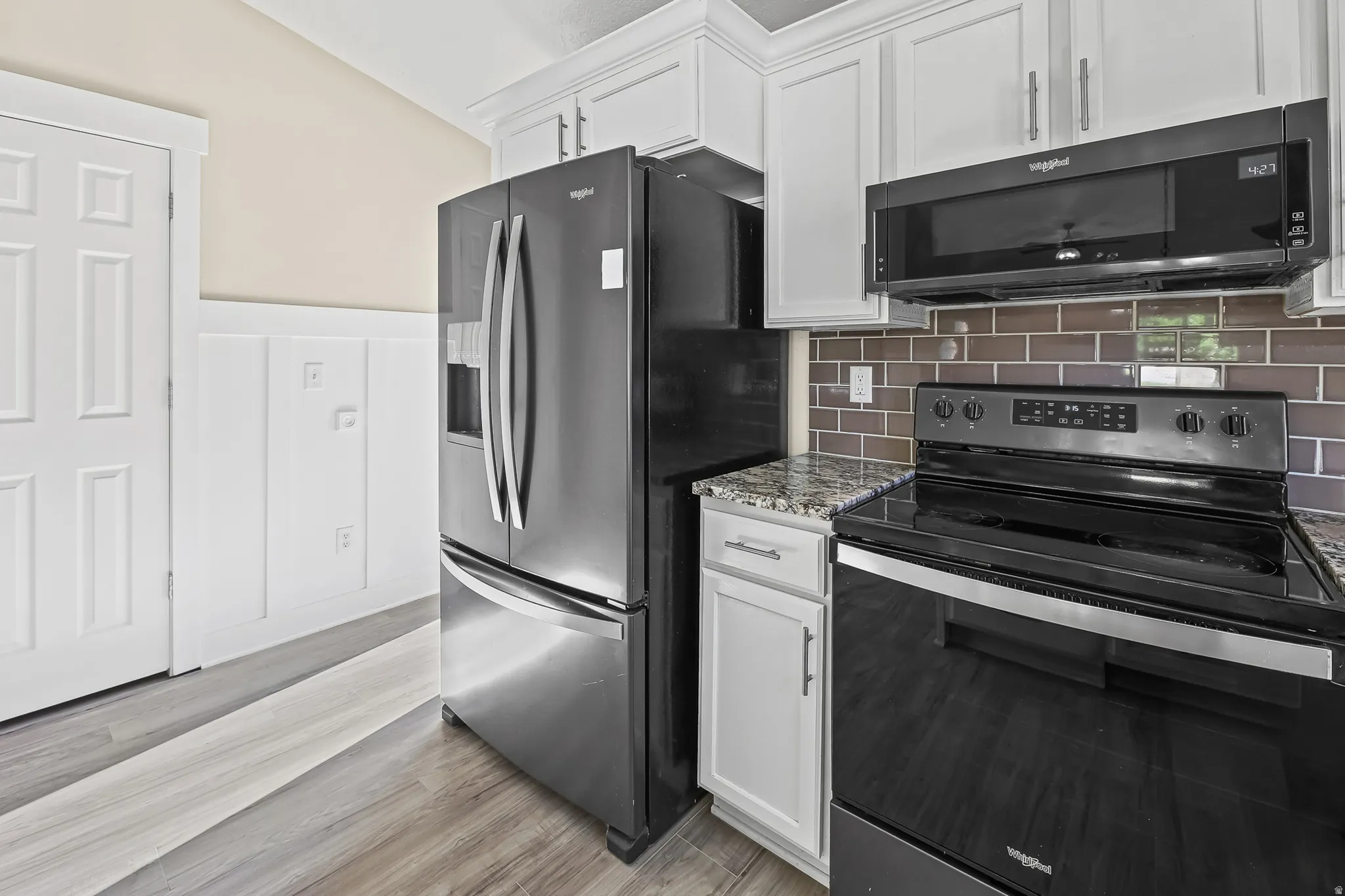 Kitchen area featuring white cabinetry, granite-style countertops, stainless steel refrigerator, black electric range with an overhead microwave, and a brown subway tile backsplash