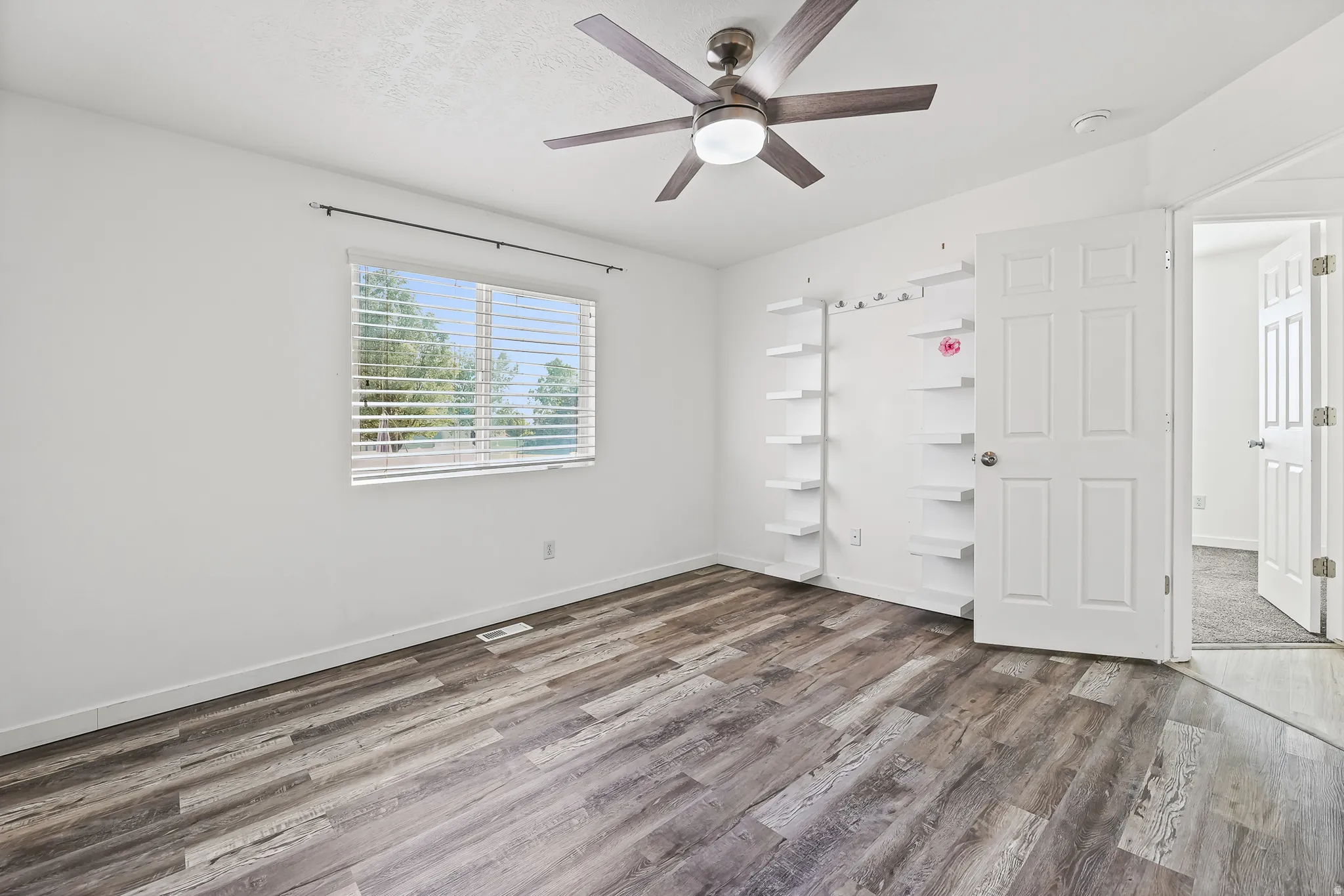 Spacious room featuring wood-finish flooring, a ceiling fan with integrated lighting, built-in shelving, and a window with horizontal blinds
