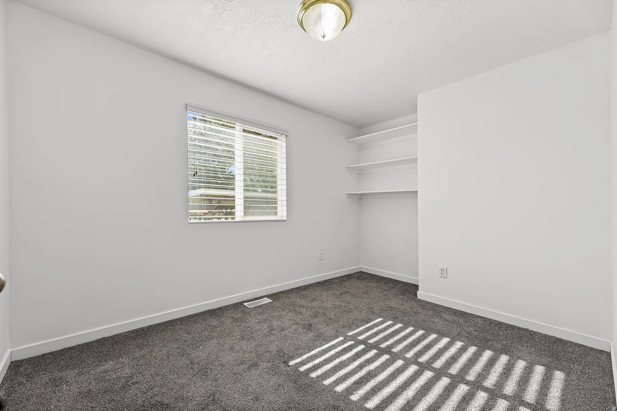 Neutral-toned room featuring a single window with blinds, carpet flooring, a flush-mount ceiling light fixture, and built-in shelving