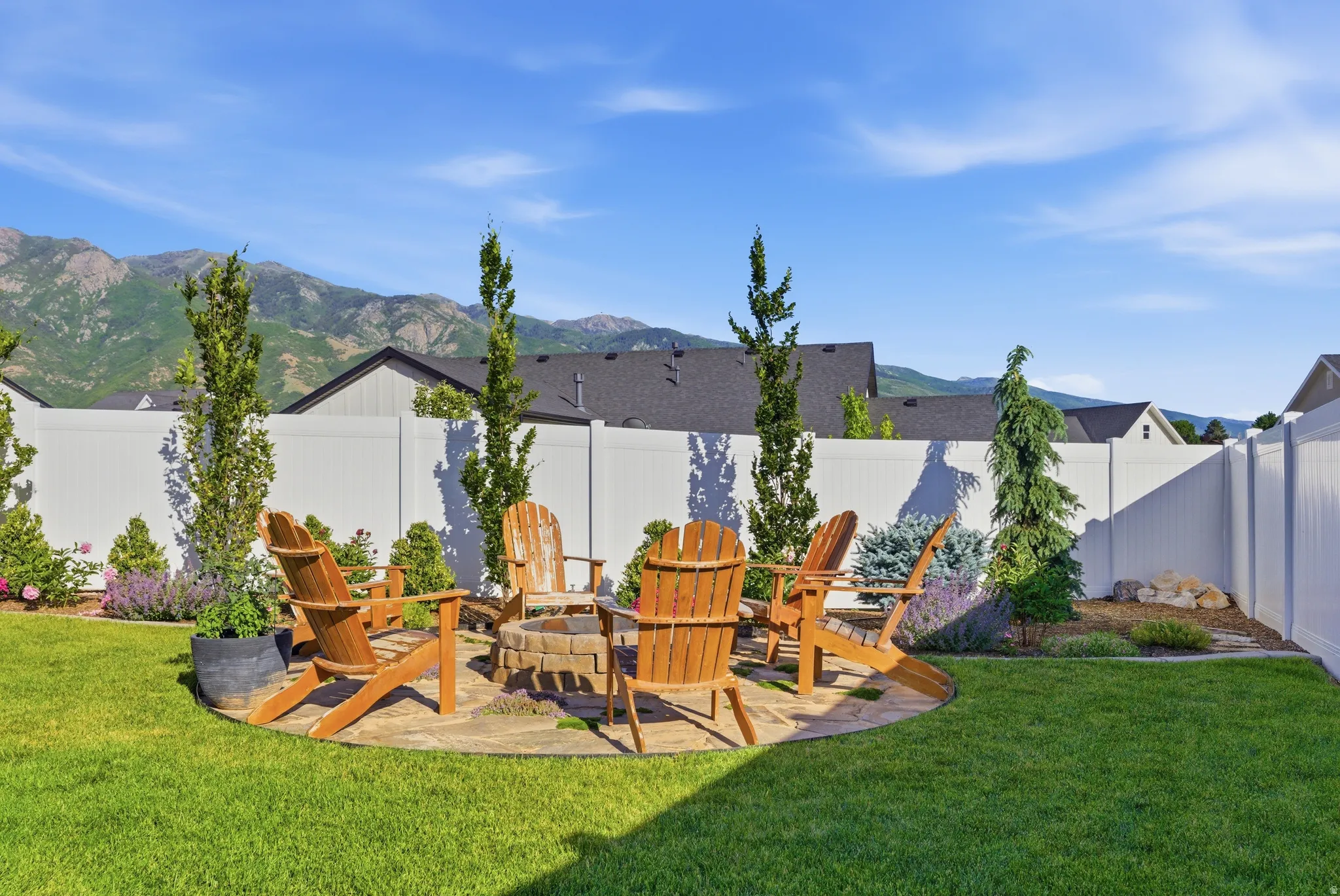 Expansive backyard featuring a circular stone patio, integrated fire pit, and surrounding garden beds with mature trees