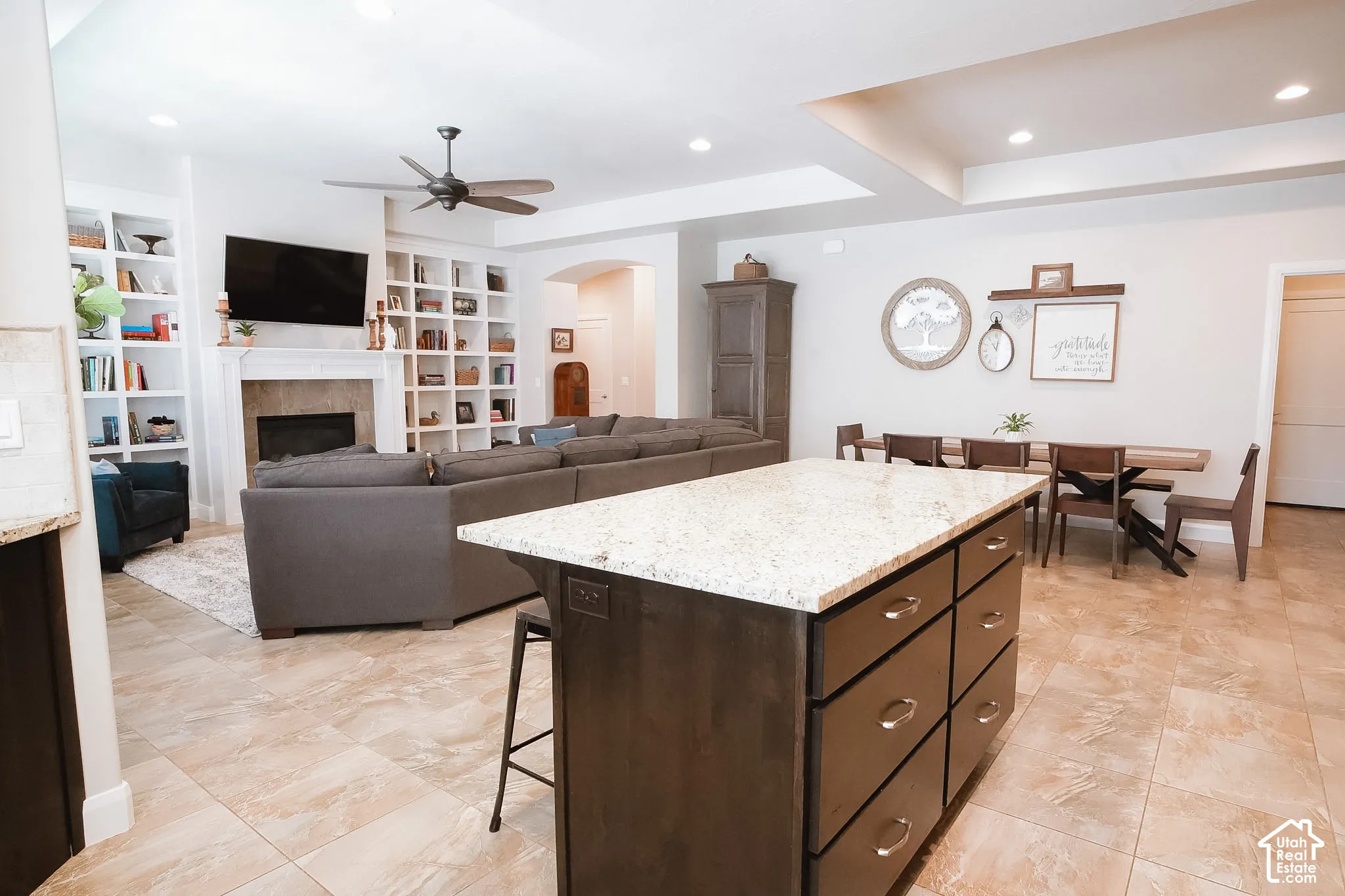 Spacious open-concept living area featuring a kitchen island with granite-style countertop, tile flooring, a fireplace with a mantel, and built-in shelving