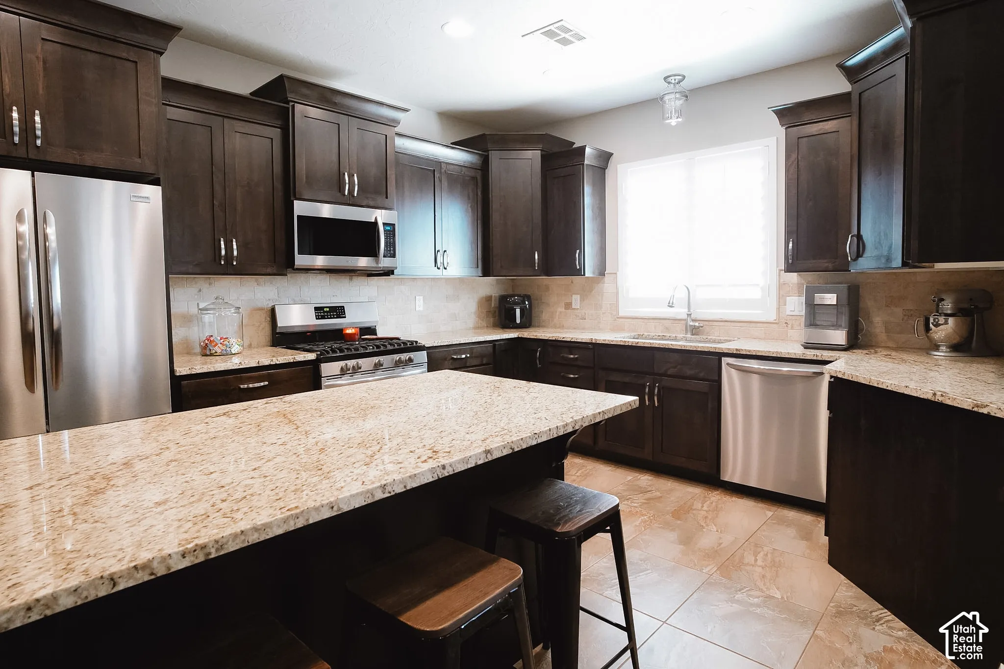 Kitchen featuring a large central island with light-toned granite countertops, dark wood cabinetry, stainless steel appliances, and a tile backsplash