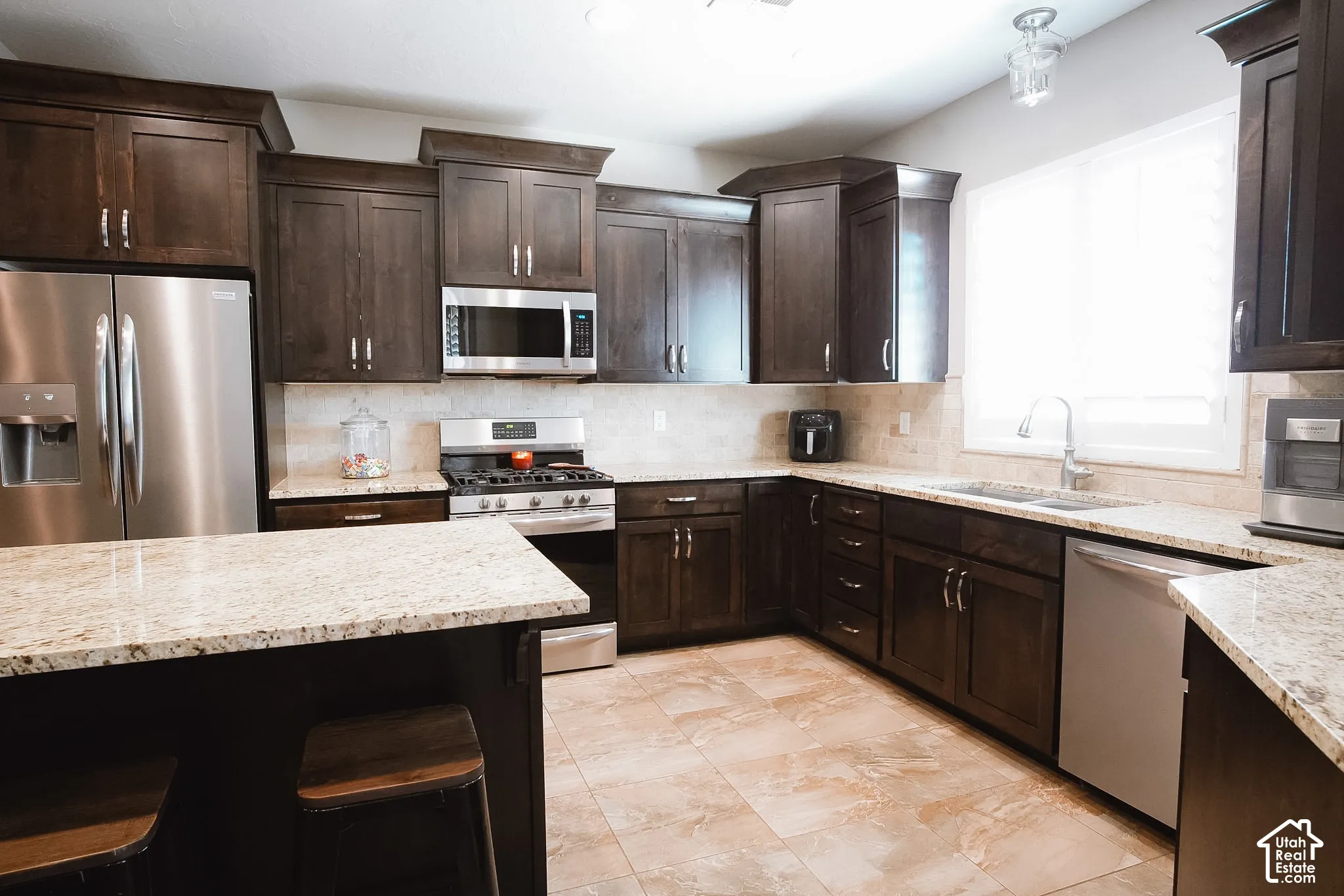 Kitchen featuring dark wood cabinetry, light-toned countertops, stainless steel appliances, a tile backsplash, and light-toned floor tiling