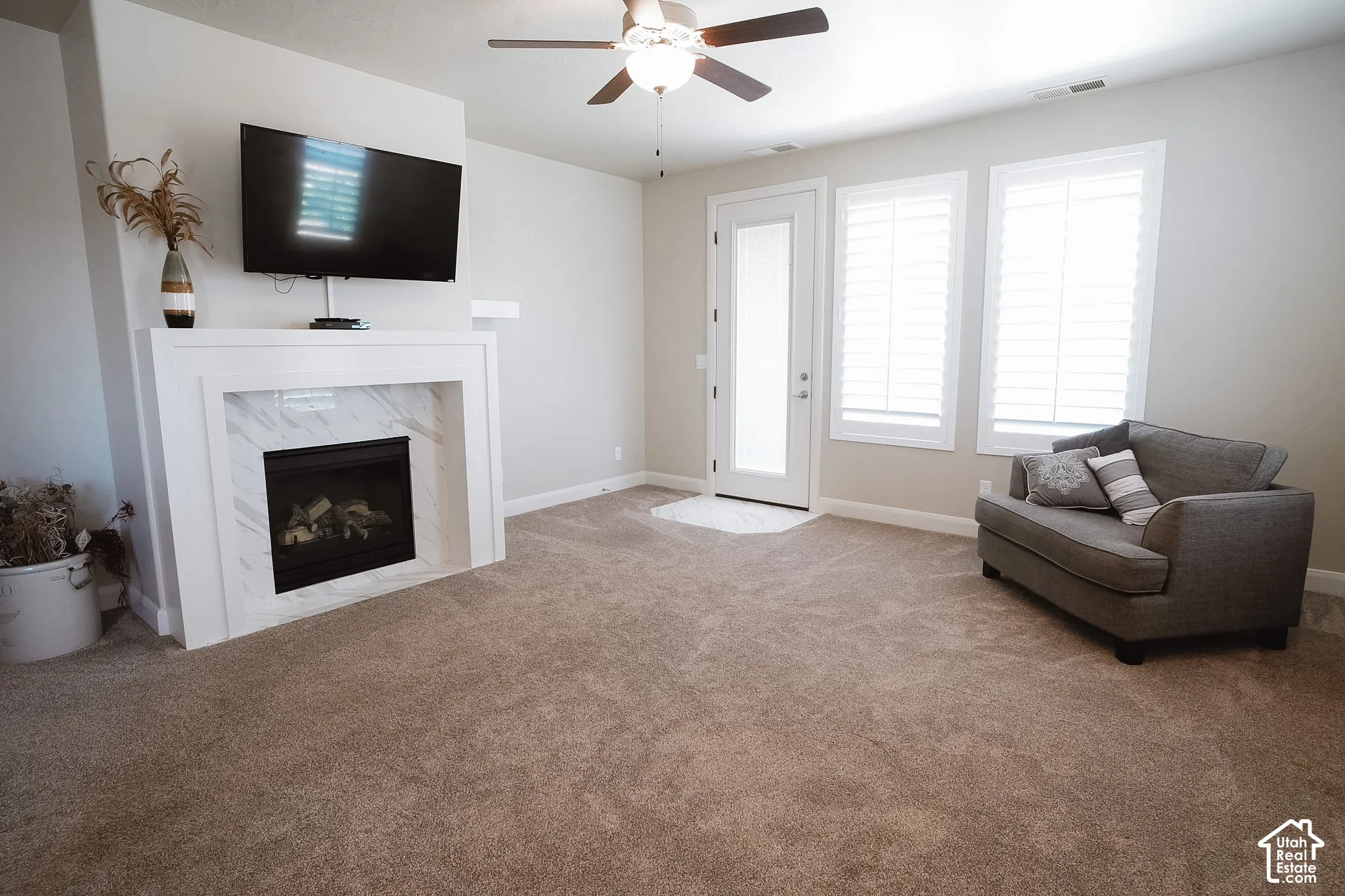 Living space featuring a white mantel fireplace with a marble-finish surround, carpet flooring, a ceiling fan, a glass-paneled door, and two windows with white plantation shutters