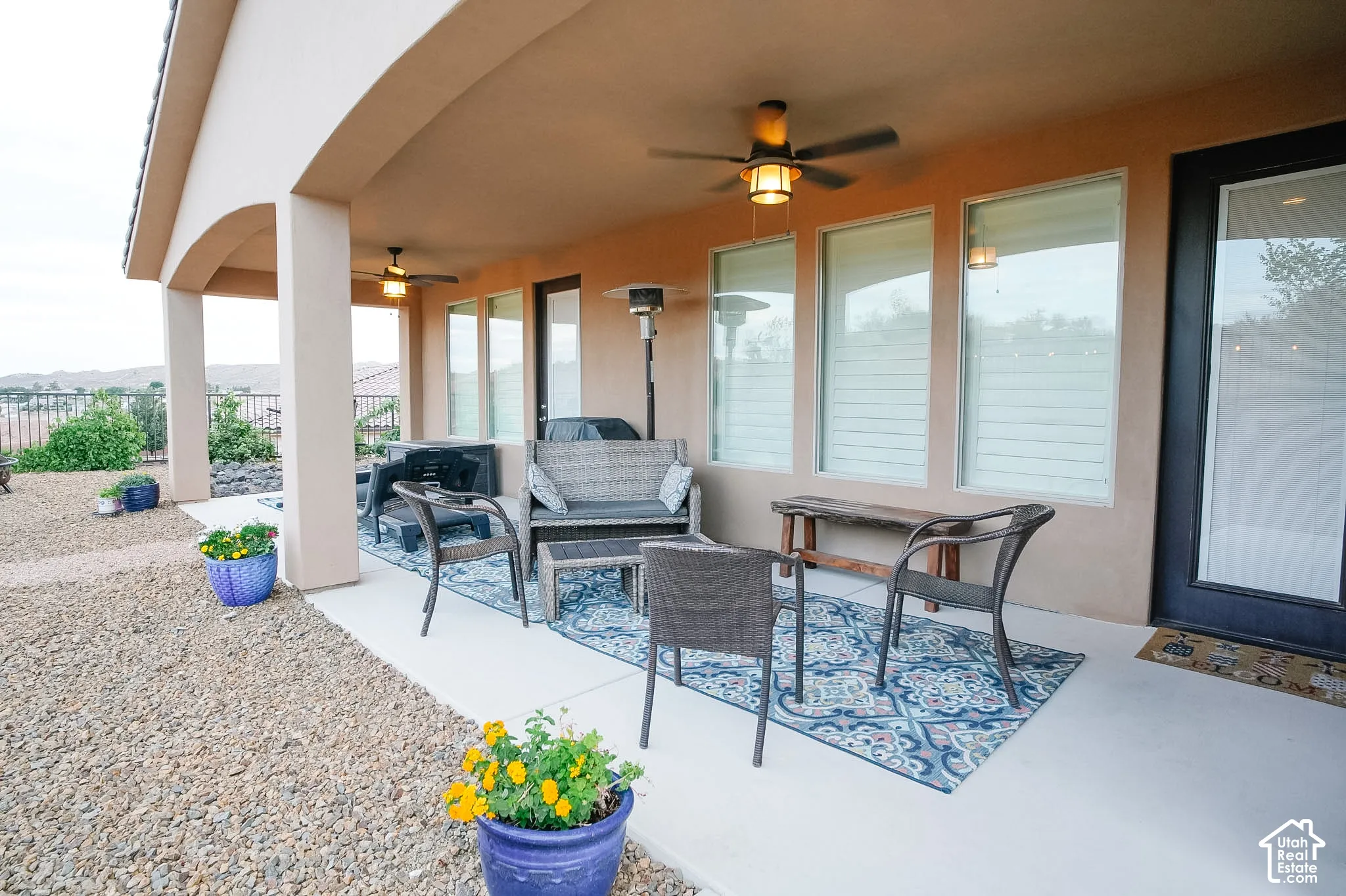 Expansive covered patio featuring two ceiling fans with integrated lighting, three large windows with privacy blinds, and a glass-paneled exterior door