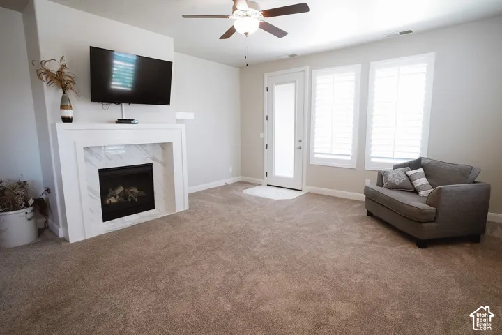 Living space featuring a white mantel fireplace with a marble-finish surround, carpet flooring, a ceiling fan, a glass-paneled door, and two windows with white plantation shutters