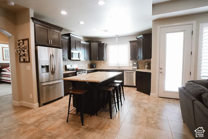 Spacious kitchen featuring dark wood cabinetry, stainless steel appliances, a large center island with seating, ample countertop space, and light-toned tile flooring
