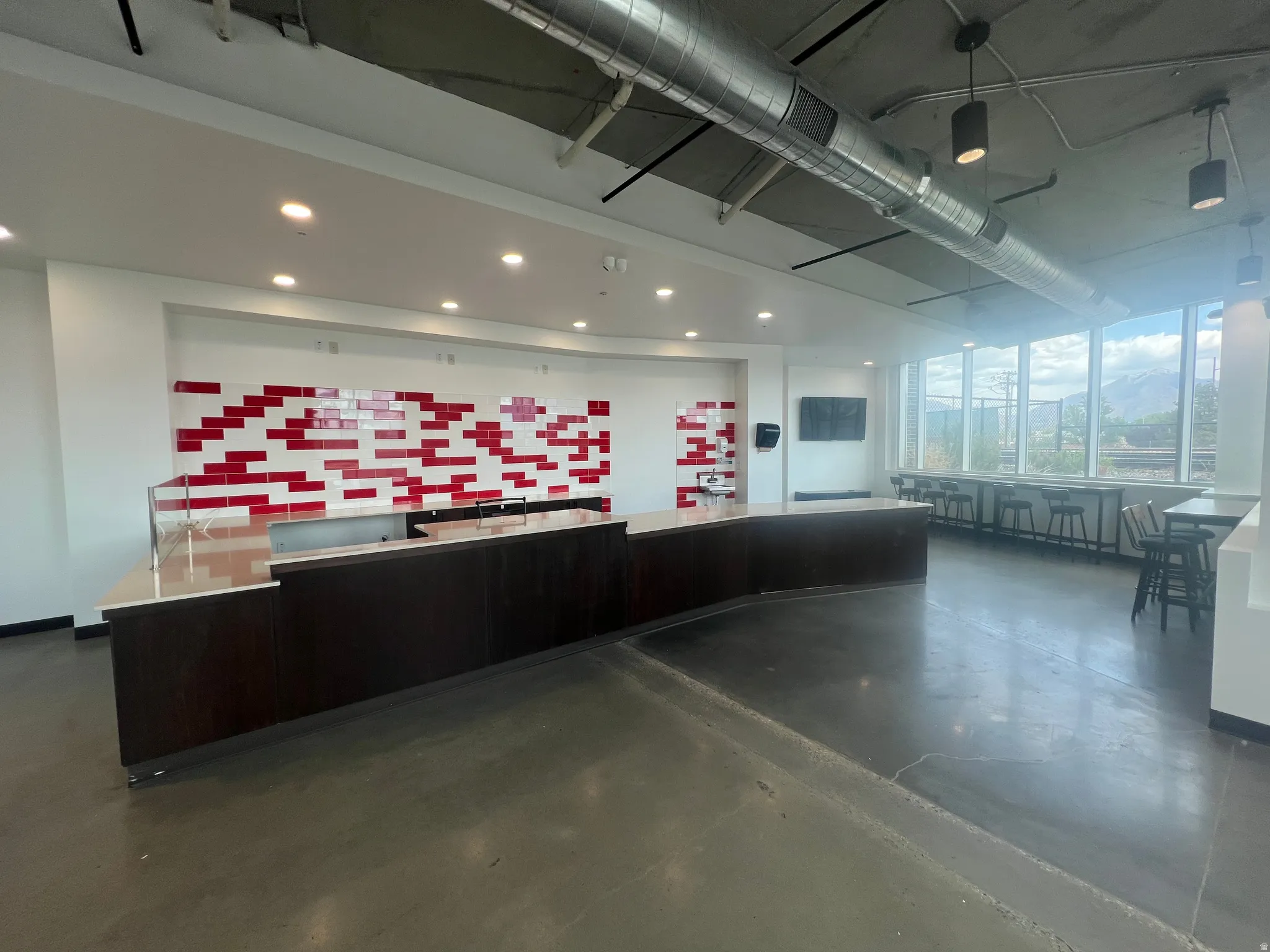 Spacious interior featuring a large serving counter with a light-toned surface, concrete flooring, exposed ceiling ductwork, and abundant natural light from extensive windows