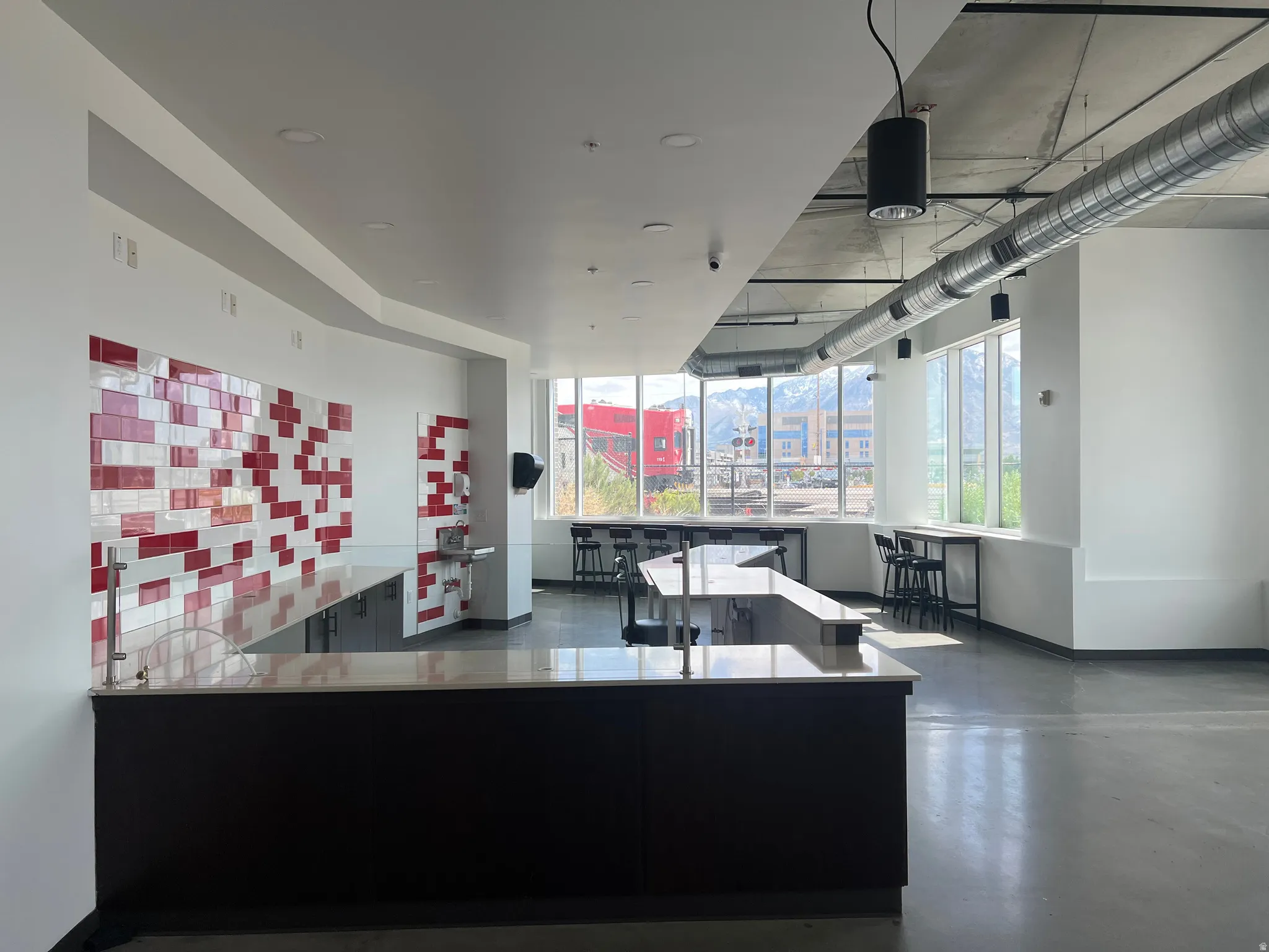 Open concept space featuring a large central island with a light-toned countertop, extensive windowline, exposed ductwork, concrete flooring, and a red and white tiled accent wall