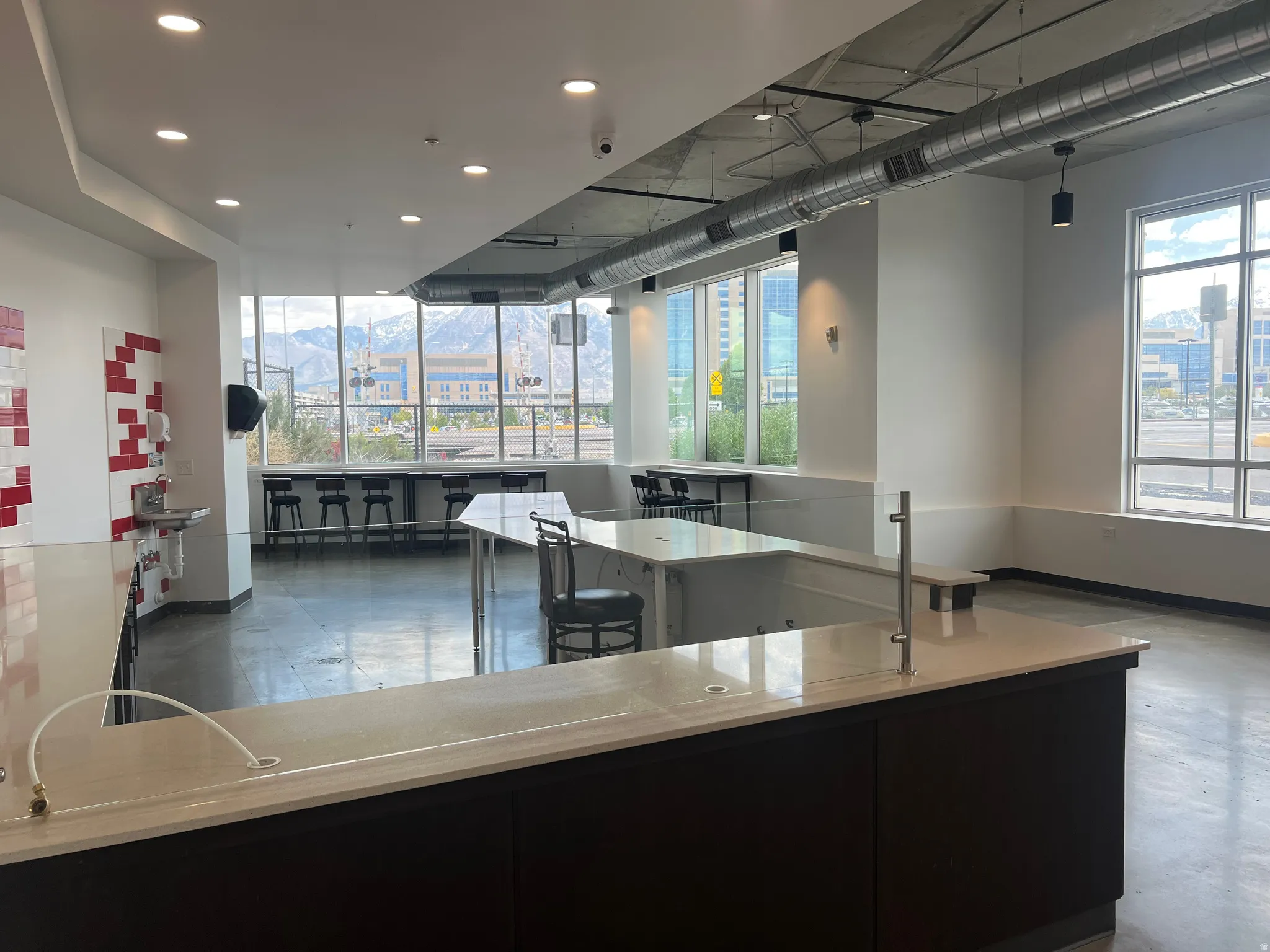 Spacious open-concept commercial interior featuring floor-to-ceiling windows, exposed industrial ductwork, polished concrete flooring, a large service counter with a light-toned countertop, and modern recessed lighting