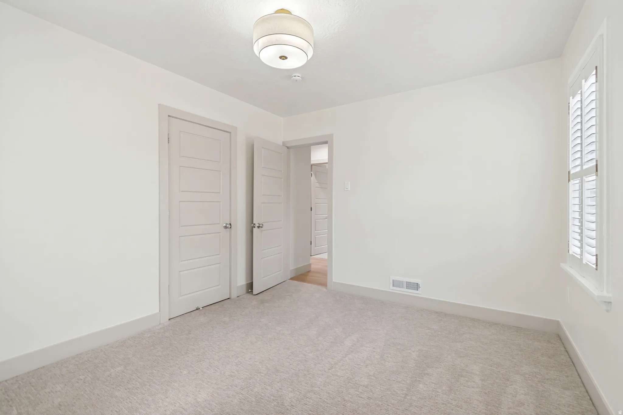 Carpeted room featuring a ceiling-mounted light fixture, white walls, and a window with plantation shutters
