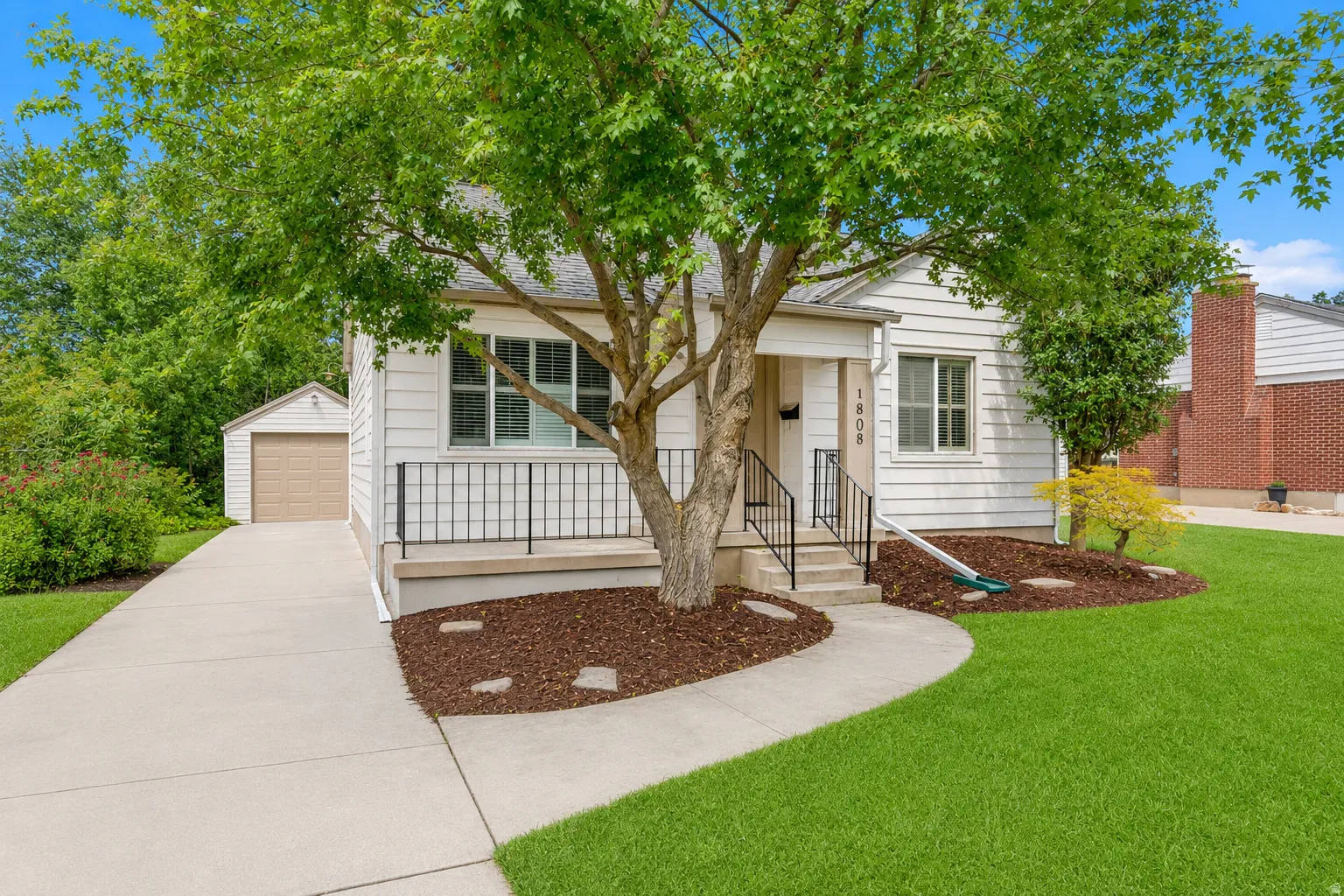 Charming white siding residence featuring a covered front porch, a well-maintained lawn, mature trees, a paved driveway, and a detached garage