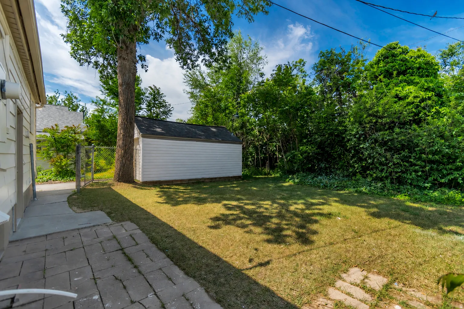 Expansive backyard featuring a white storage shed with dark roofing, a mature tree providing natural shade, a partial chain-link fence, and a stone paver patio