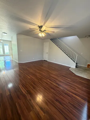 Spacious living area featuring rich wood-finish flooring, a contemporary ceiling fan with integrated lighting, and a white staircase with open railings