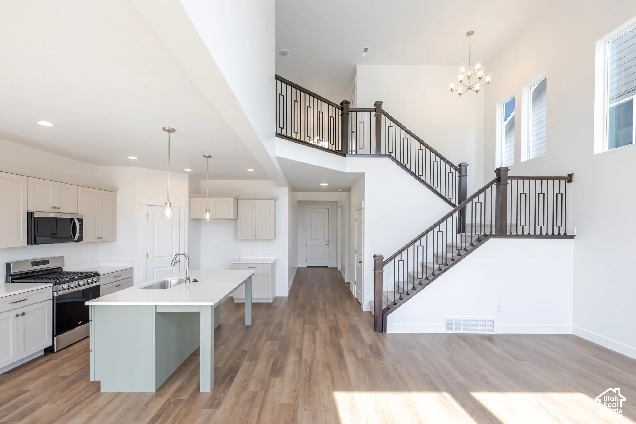 Two-story ceiling with large windows, wood-finish flooring, staircase with wood and metal railing, kitchen island with sink, and stainless steel appliances