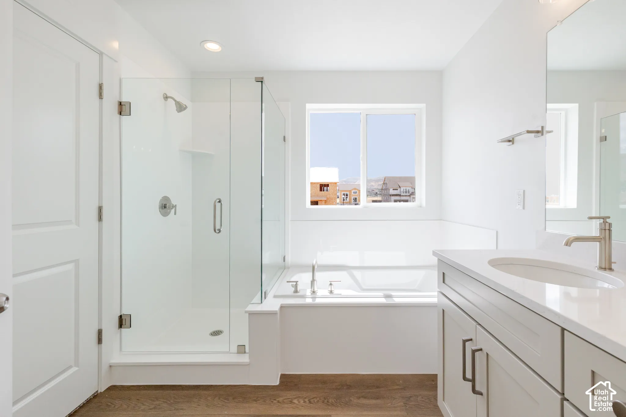 Spacious bathroom featuring a glass-enclosed shower, built-in bathtub, dual-pane window, single vanity with an integrated sink, and wood-finish flooring