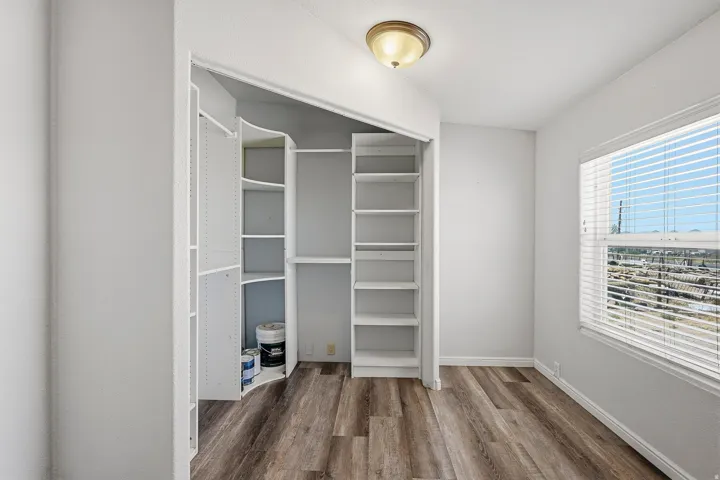 Interior room featuring a built-in closet system with adjustable shelving, wood-finish flooring, a ceiling-mounted light fixture, and a window with horizontal blinds