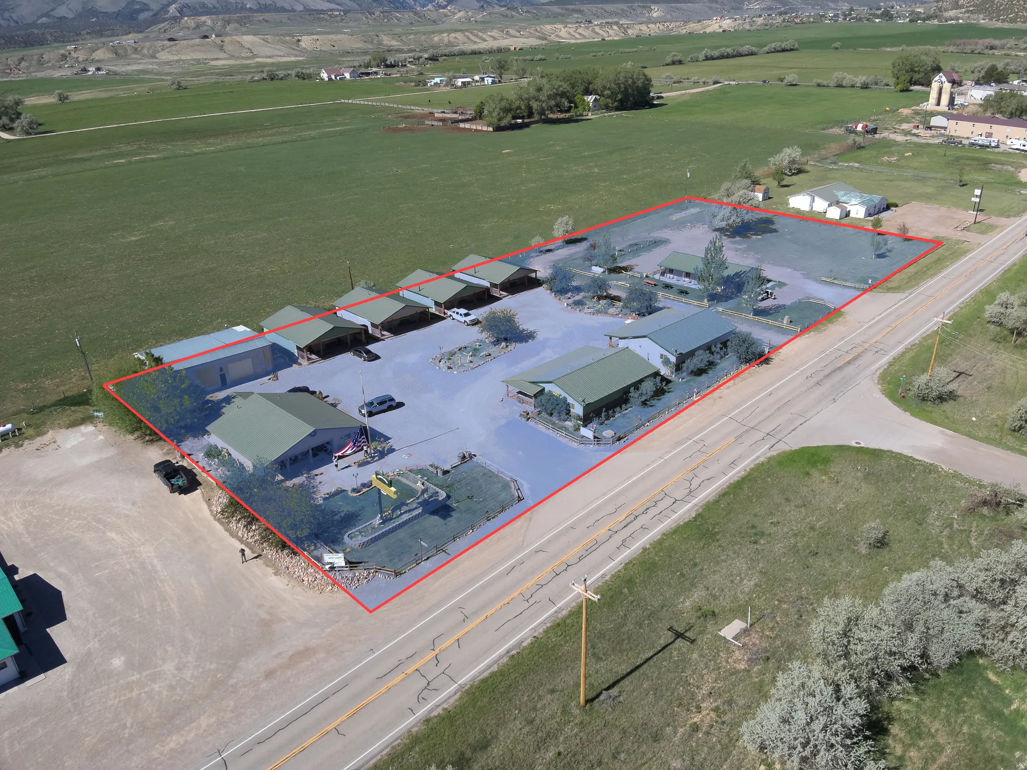Expansive commercial property featuring multiple buildings with green metal roofs, paved parking areas, mature trees, and direct road frontage