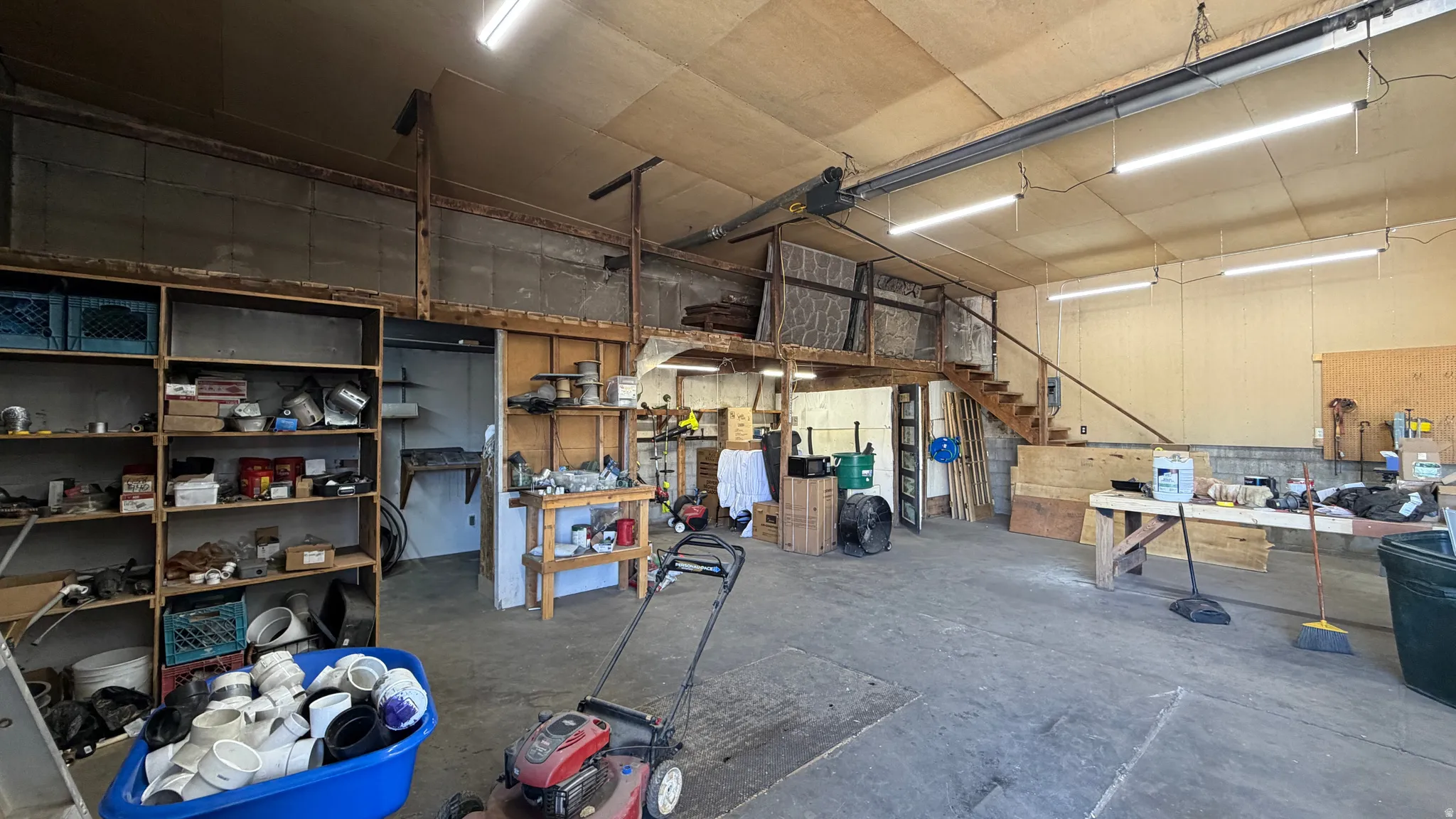 Spacious workshop featuring concrete flooring, an overhead garage door track, an integrated wooden staircase, a built-in wooden workbench, and overhead fluorescent lighting