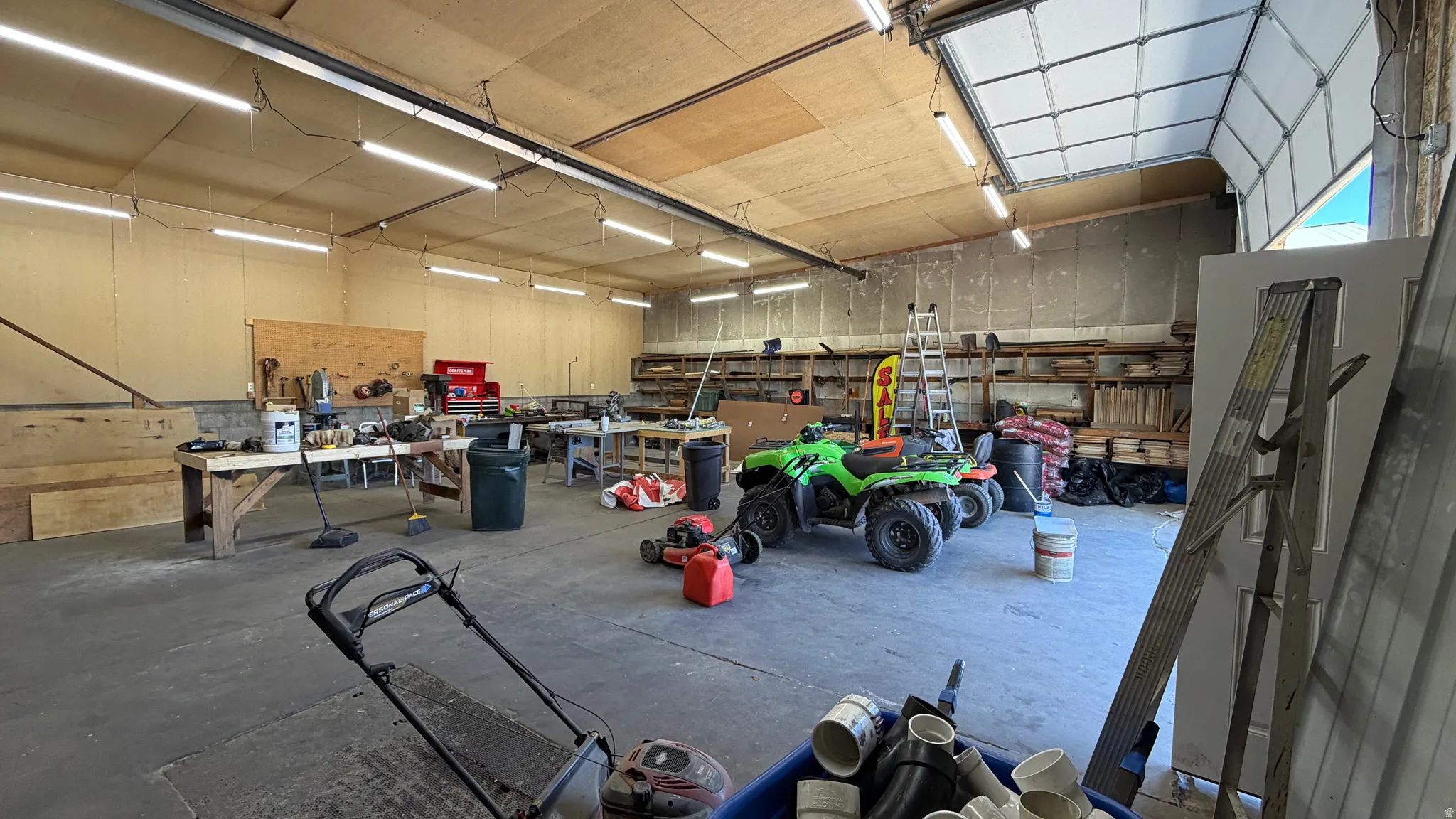 Spacious multi-purpose room featuring a large overhead door, multiple fluorescent light fixtures, exposed wood ceiling, multiple workbenches, and open shelving