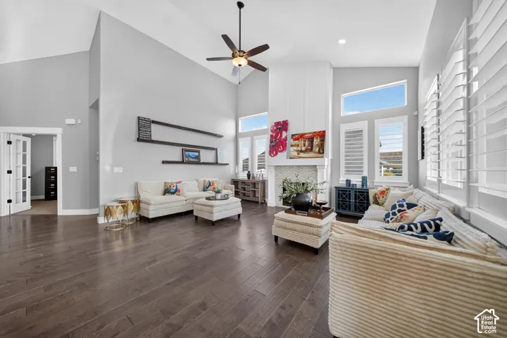Spacious open-concept living area showcasing soaring ceilings with ceiling fan, abundant natural light from large transom and picture windows with plantation shutters, dark wood-finish flooring, and neutral wall finishes