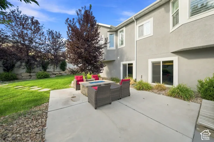 Expansive backyard featuring a concrete patio, built-in fire pit, manicured lawn with stepping stones, and mature trees