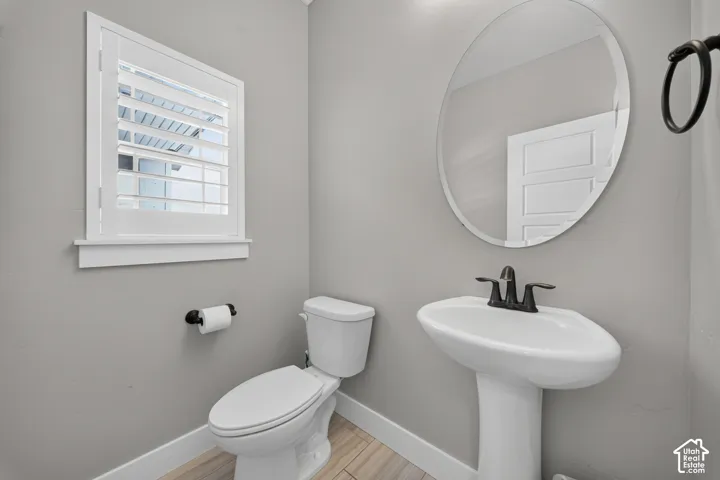 Bright powder room featuring a pedestal sink with matte black faucet, white ceramic toilet with elongated bowl, circular mirror with metal frame, wood-finish flooring, and a window with plantation shutters providing natural light