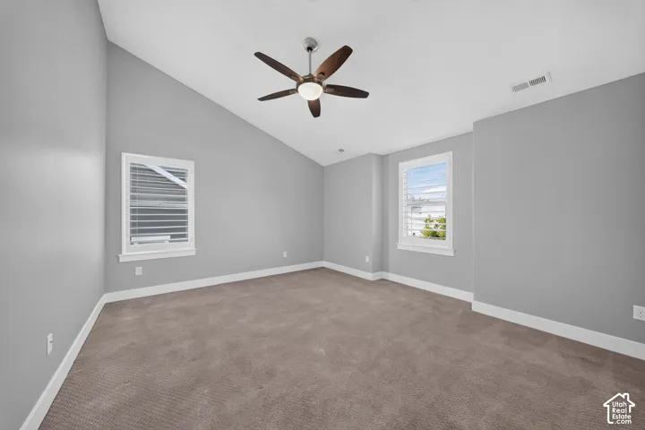 Spacious bedroom flooded with natural light from dual windows, featuring neutral gray walls, plush carpet flooring, and a modern ceiling fan with brushed metal and wood-finish blades