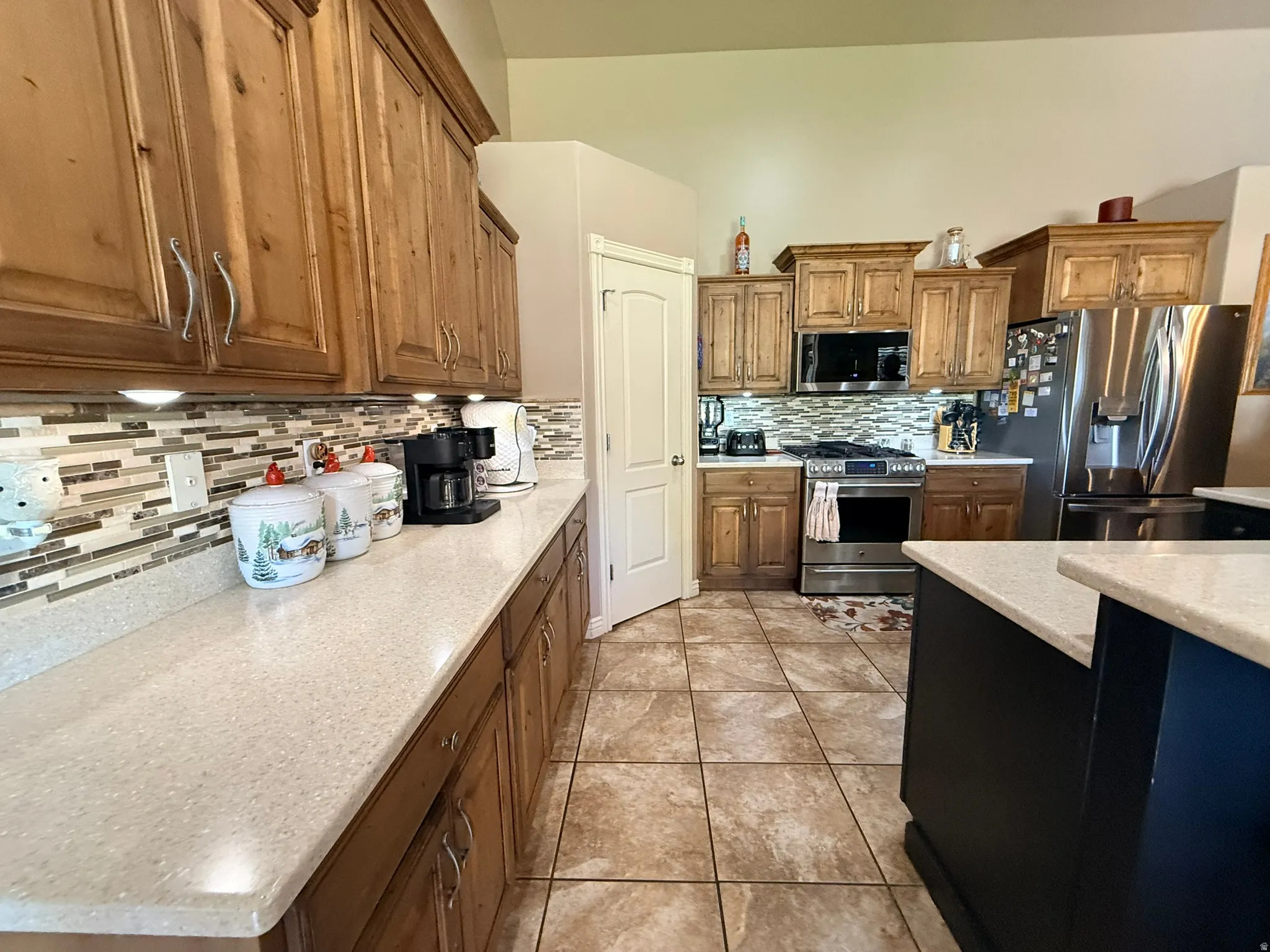 Kitchen featuring wood cabinetry, light-toned countertops, a glass tile backsplash, stainless steel appliances, and tiled flooring