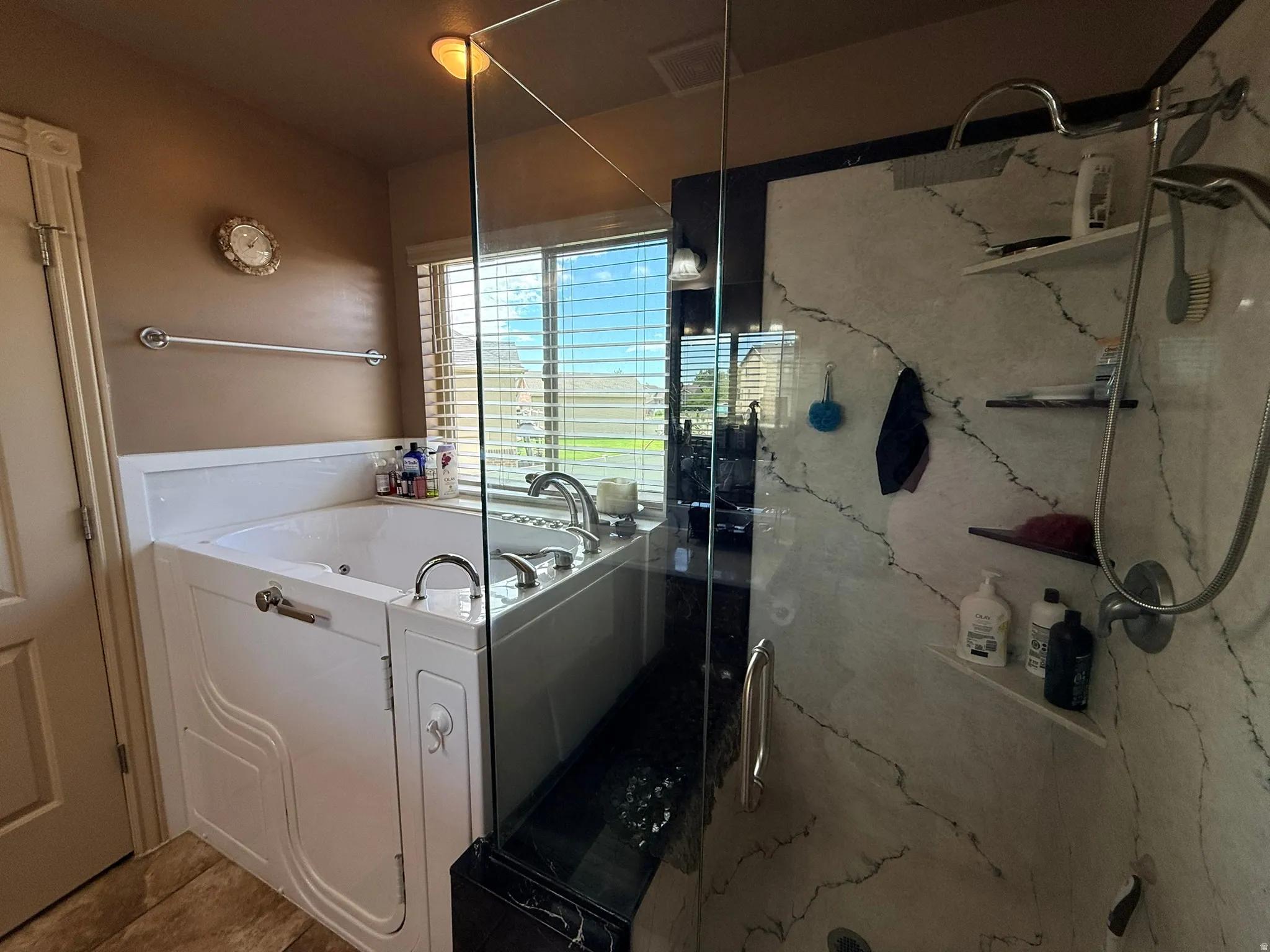 Walk-in tub with chrome fixtures, accompanied by a glass-enclosed shower featuring white and gray veined wall panels, a rainfall showerhead, and integrated shelving