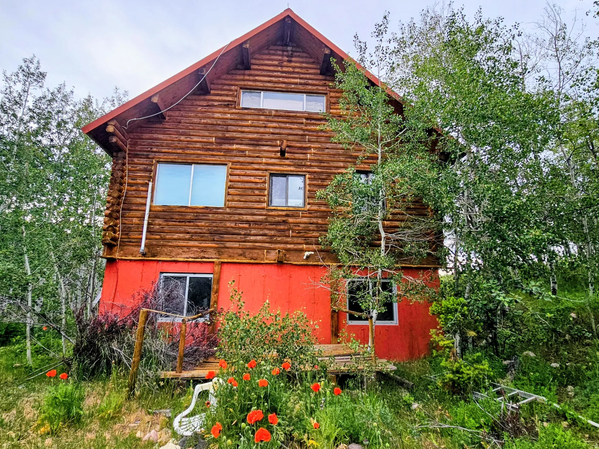 Log cabin exterior featuring a vibrant red lower level, multiple windows, and natural landscaping with mature trees and ground cover