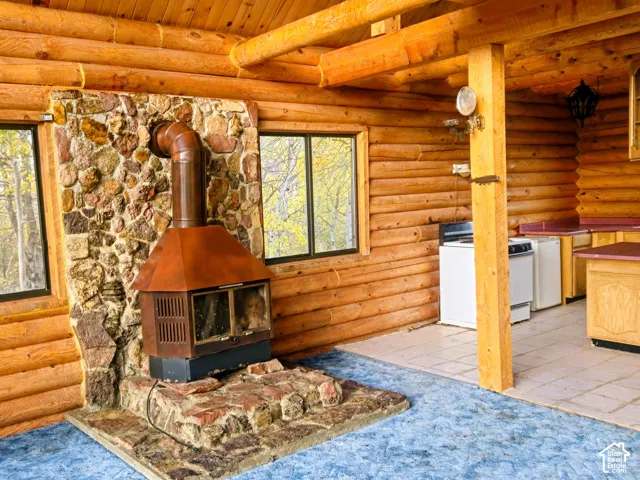 Wood log walls, stone hearth with a wood-burning stove, two windows, and light wood cabinetry