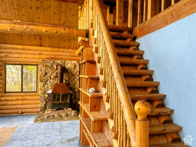 Log cabin interior featuring a stone-clad fireplace with a metal hood, wood-plank walls and ceiling, a wood-finish staircase with turned balusters, and a light-colored wall texture