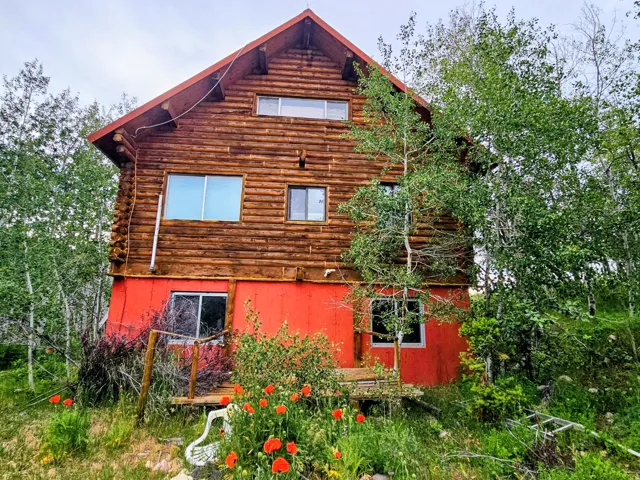 Log cabin exterior featuring a vibrant red lower level, multiple windows, and natural landscaping with mature trees and ground cover