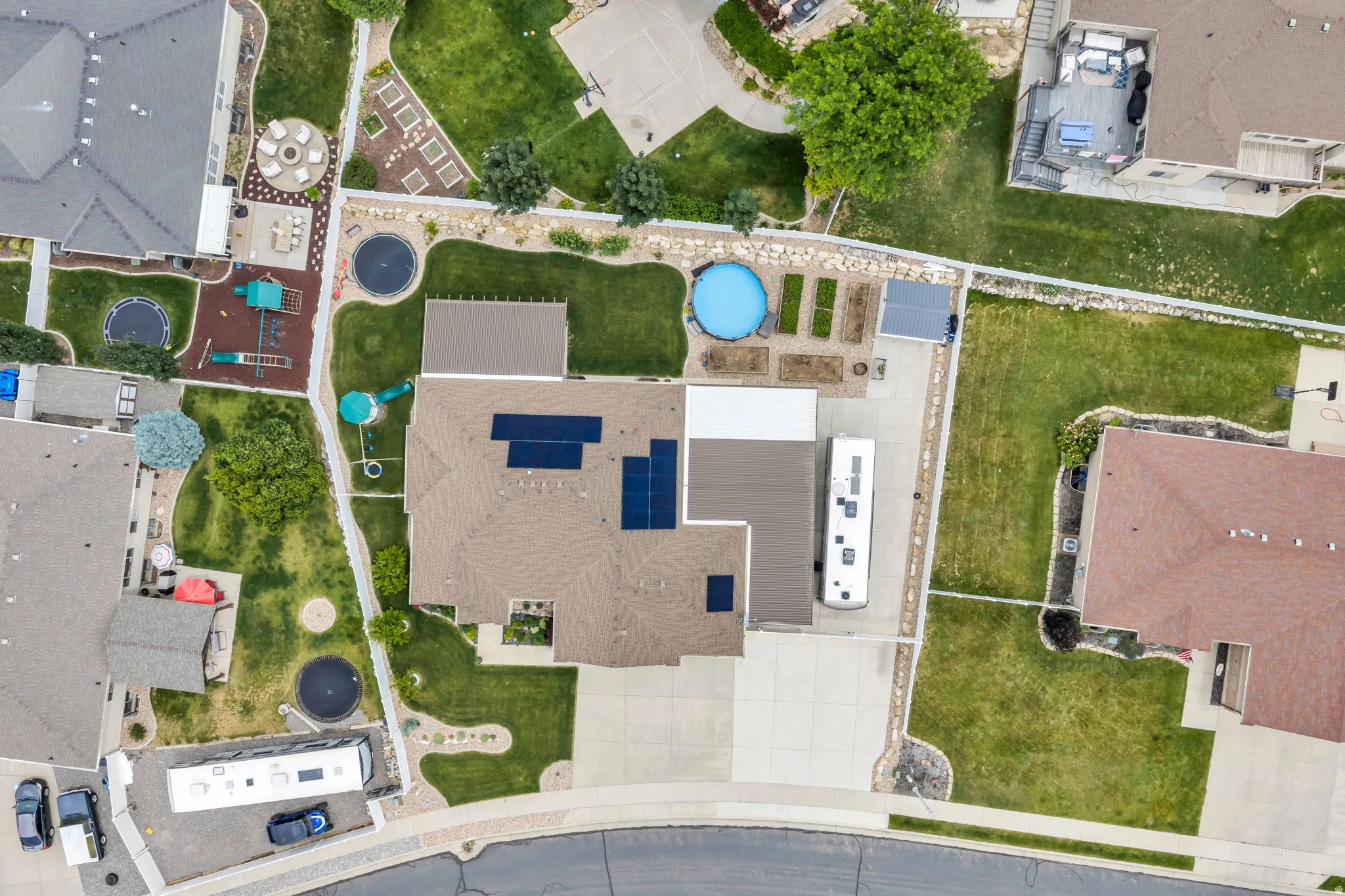 Aerial property view featuring a brown shingle roof with solar panels, a concrete driveway with RV parking, a backyard with an above-ground pool, and garden beds