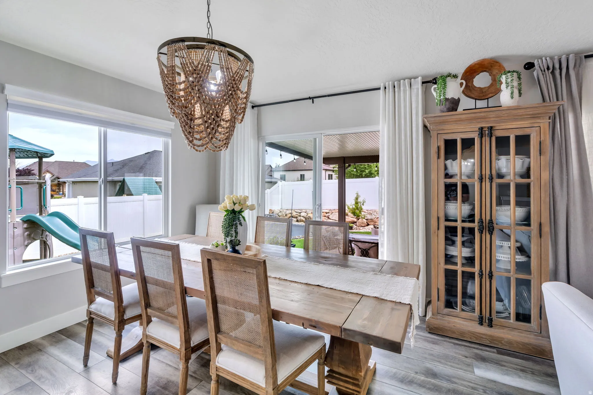 Spacious dining area featuring wood-finish flooring, a contemporary beaded chandelier, and abundant natural light from a large window and sliding glass door
