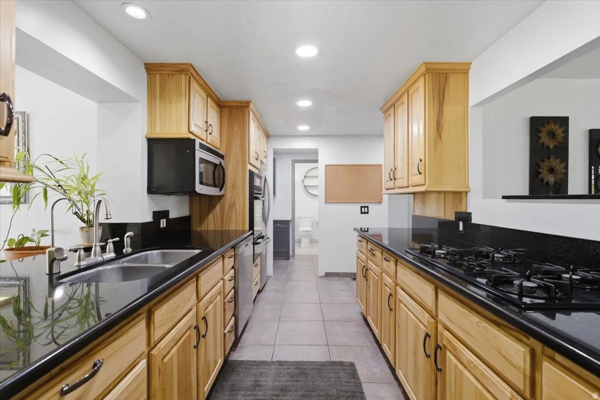 Galley kitchen featuring light wood cabinetry with dark hardware, black countertops, stainless steel appliances, and recessed lighting