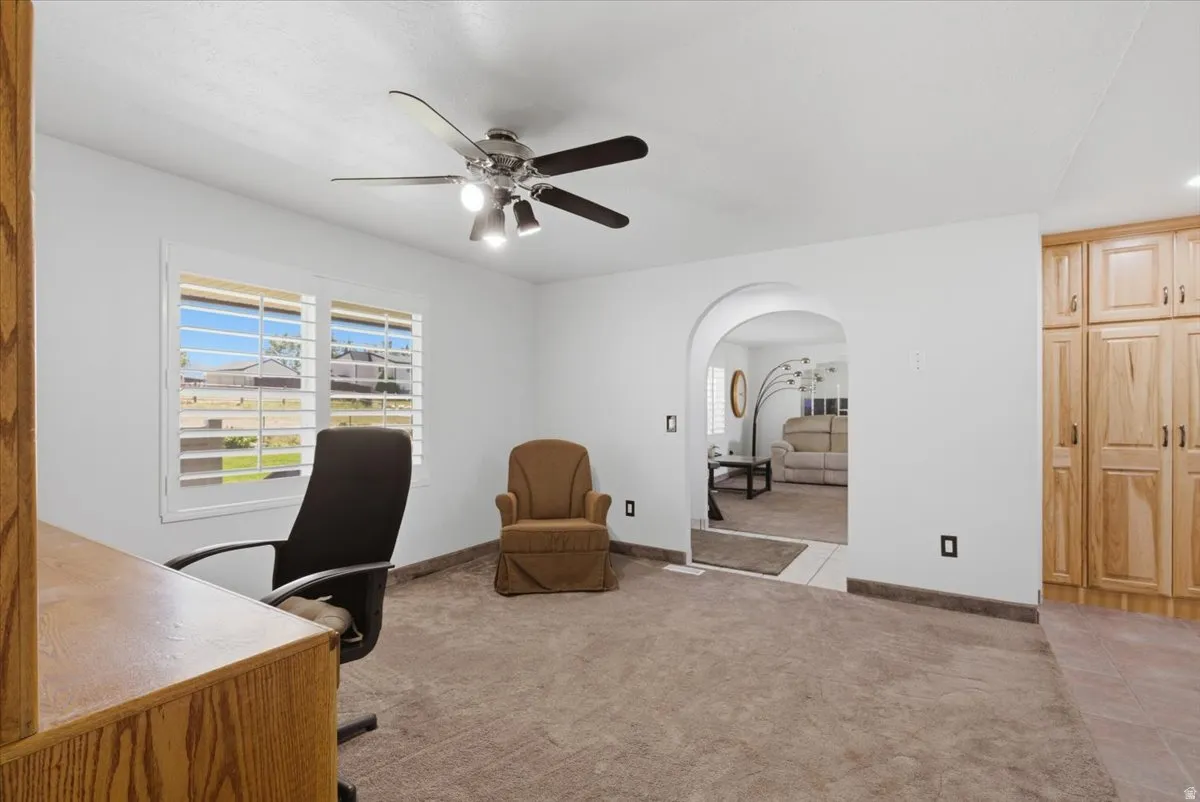 Spacious room featuring carpeted flooring, a window with plantation shutters, ceiling fan with integrated lighting, and an arched entryway to an adjoining room