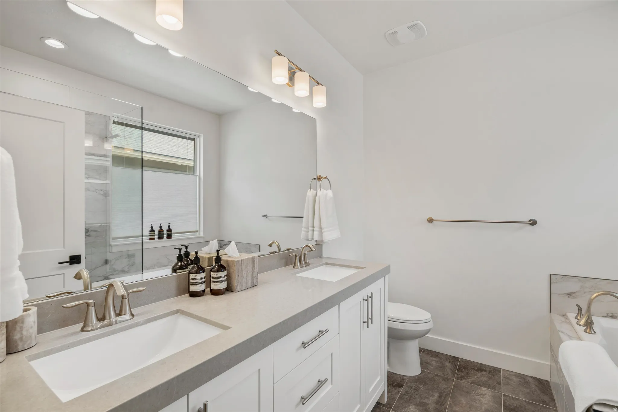 Bathroom featuring a double vanity with light gray countertops and undermount sinks, white cabinetry with brushed nickel hardware, and a large mirror with integrated lighting