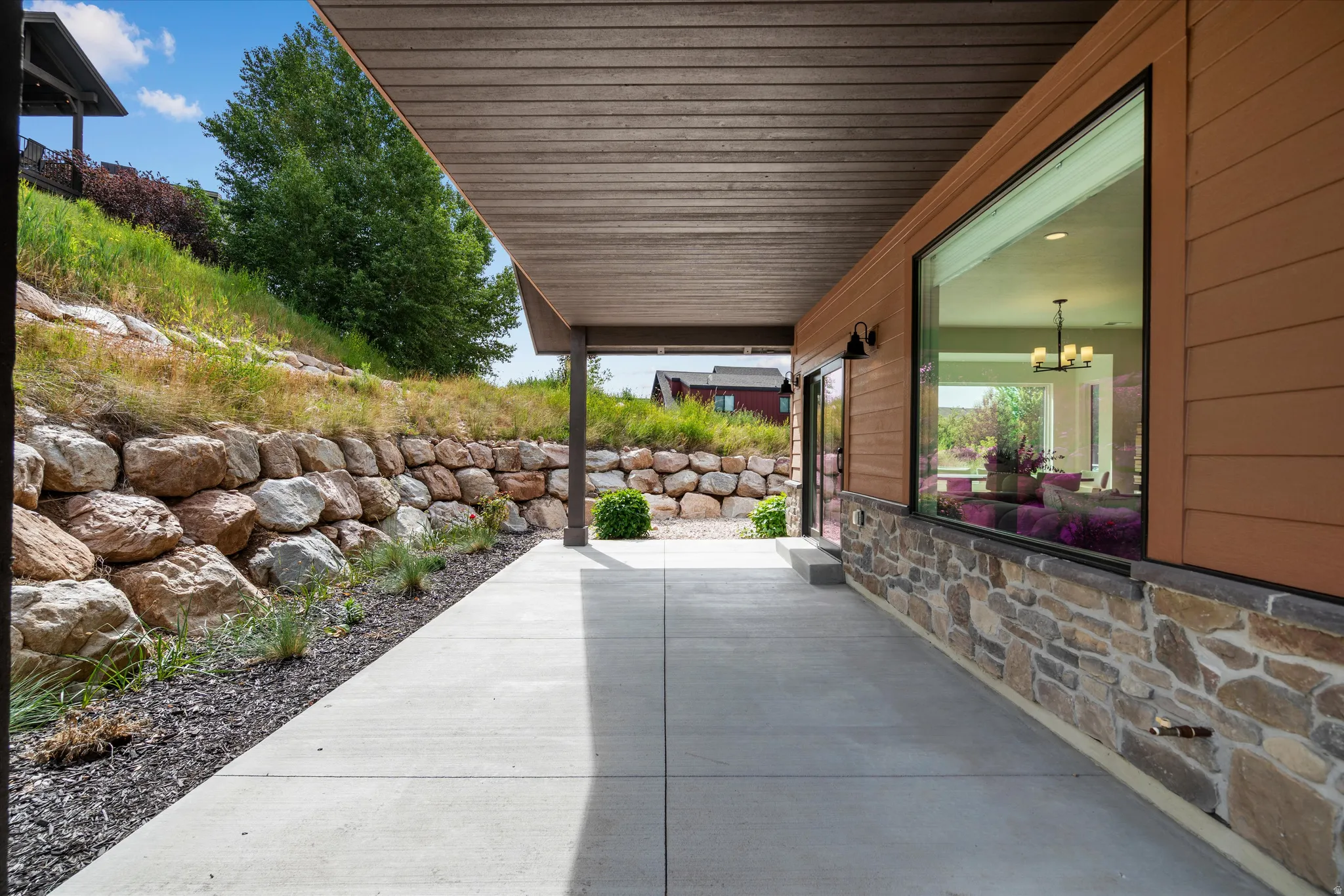Covered concrete patio with wood-finish ceiling, flanked by a stacked stone retaining wall and a horizontal plank exterior.