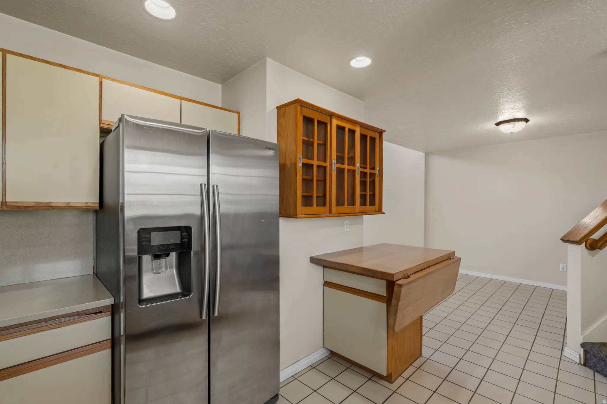 Kitchen area featuring a stainless steel side-by-side refrigerator, cream-colored upper and lower cabinetry, and a wood-finish drop-leaf counter