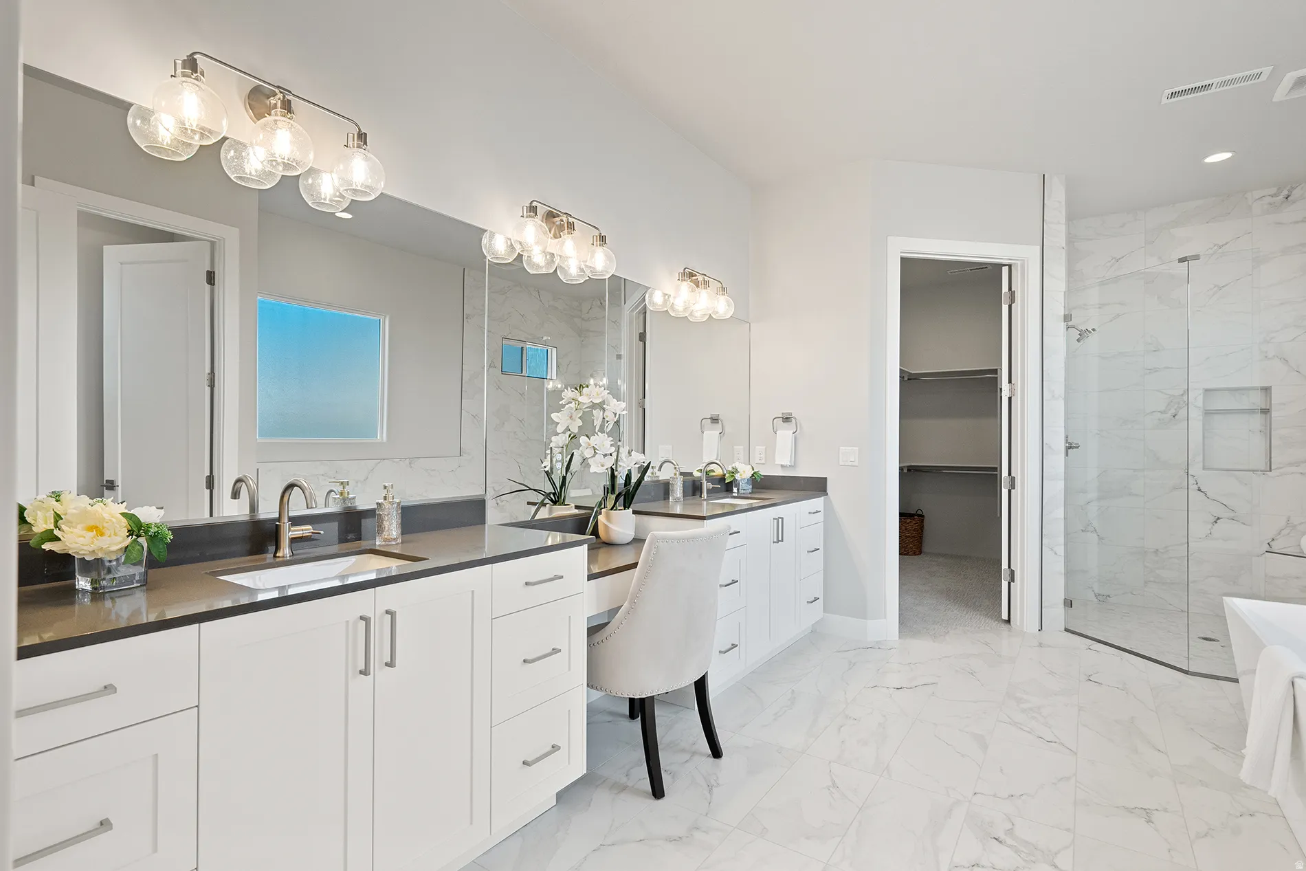 Dual vanity with undermount sinks and dark countertops, extensive marble-look tile flooring, frameless glass shower enclosure, built-in shower niche, and an open doorway to a walk-in closet