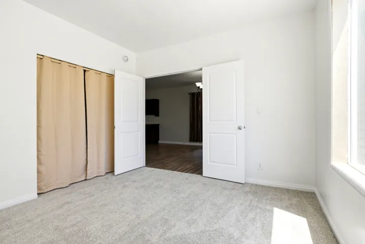 Carpeted room with white walls, featuring a window, bi-fold panel doors, and two-panel interior doors opening to an adjacent space with wood-finish flooring