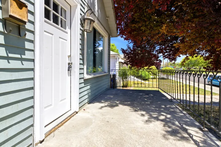 Front porch featuring a white paneled entry door, light blue siding, decorative black wrought iron railing, and a mature tree providing natural shade